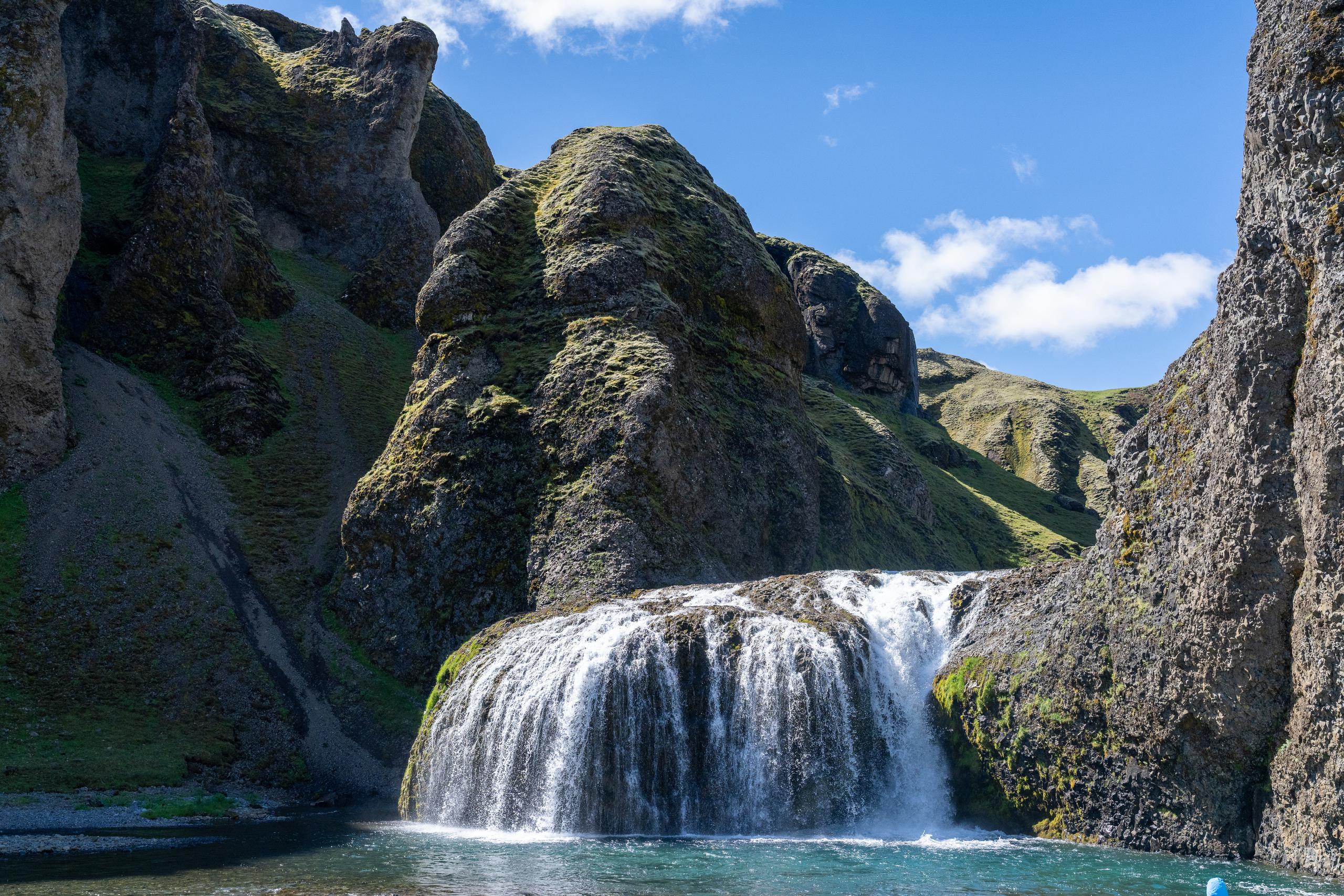 A wide waterfall pours down from the moss-covered black rock wall. The water divides into multiple streams and falls into the blue pool below. Water mist rises on the surface of the pool. On both sides of the waterfall are towering rock walls and steep slopes covered with green moss. There is also a small waterfall on the right rock wall, and a corner of blue sky and white clouds are exposed at the top of the frame.