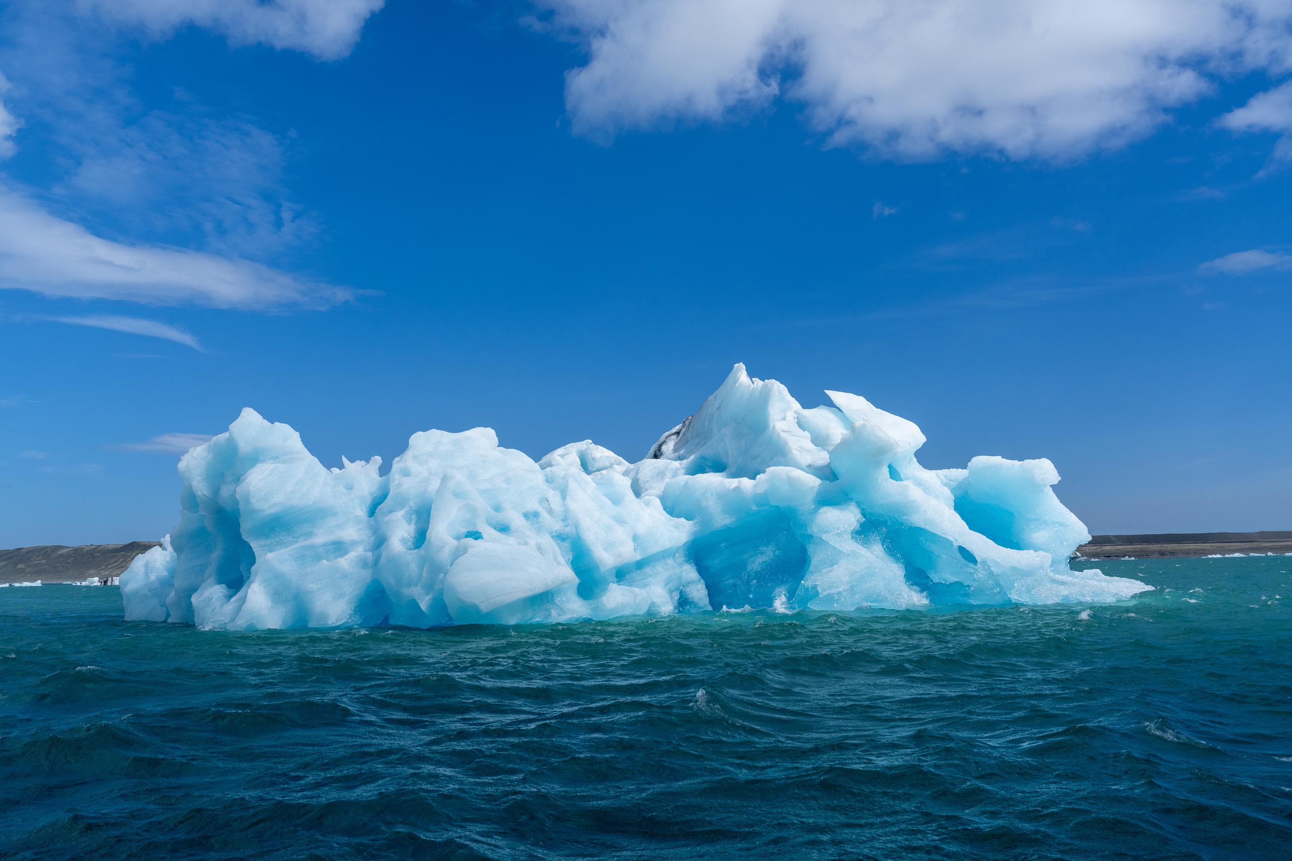 A huge iceberg floats on the dark blue sea. The ice body is white and translucent light blue, and the surface is covered with sharp corners, cracks and melted textures. The sky is dark blue, with white clouds floating in it. In the distance, there are low brown coastlines on both sides of the horizon.