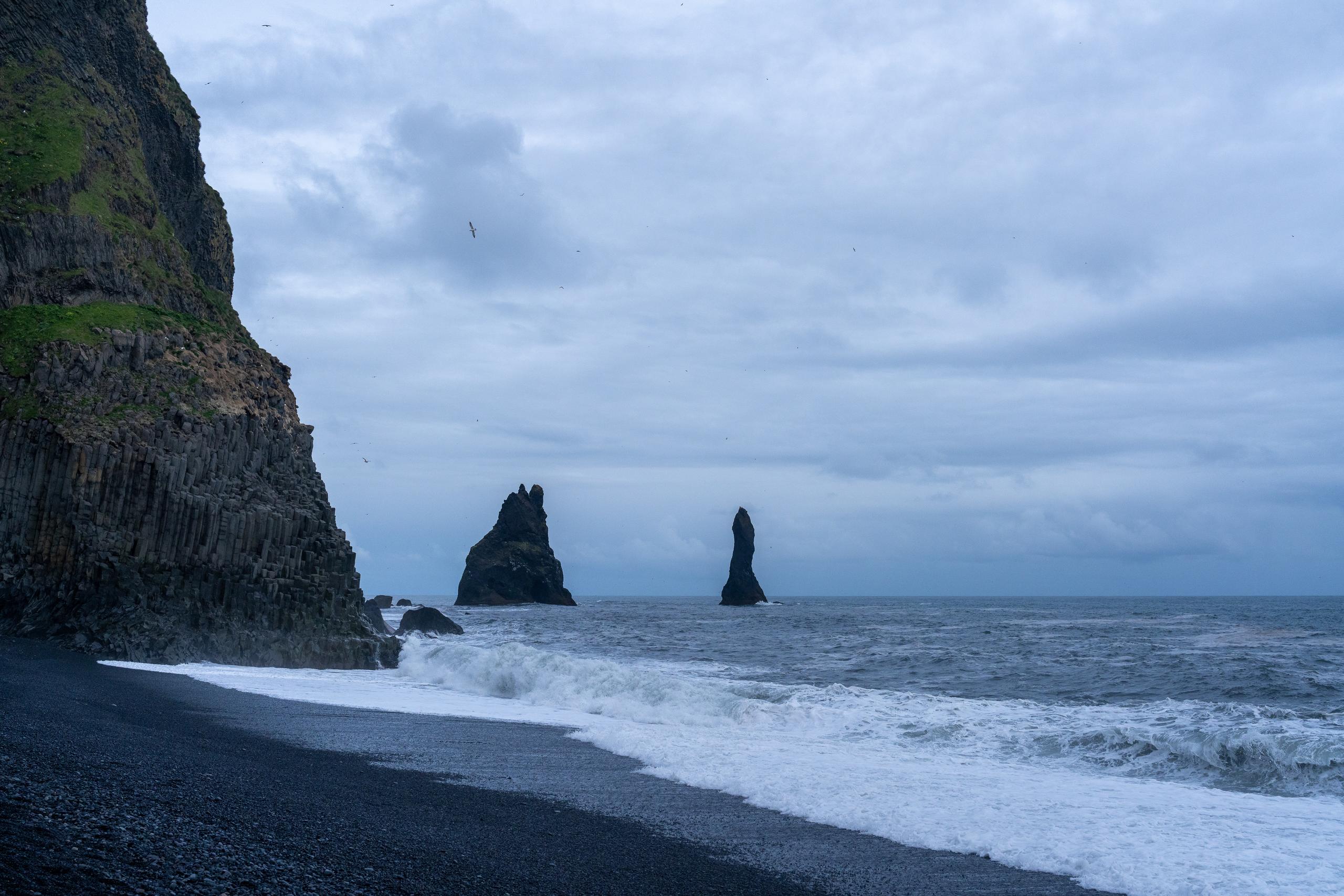 Black sand beach under a cloudy sky: in the foreground is a black gravel beach, with a white wave surging ashore; on the left is a dark rock wall higher than the frame, with green vegetation hanging on the wall; two huge black reef pillars stand on the sea, one of which is tall and pointed. In the distance, the horizon is connected with gray clouds, and there are several seabirds in the sky.