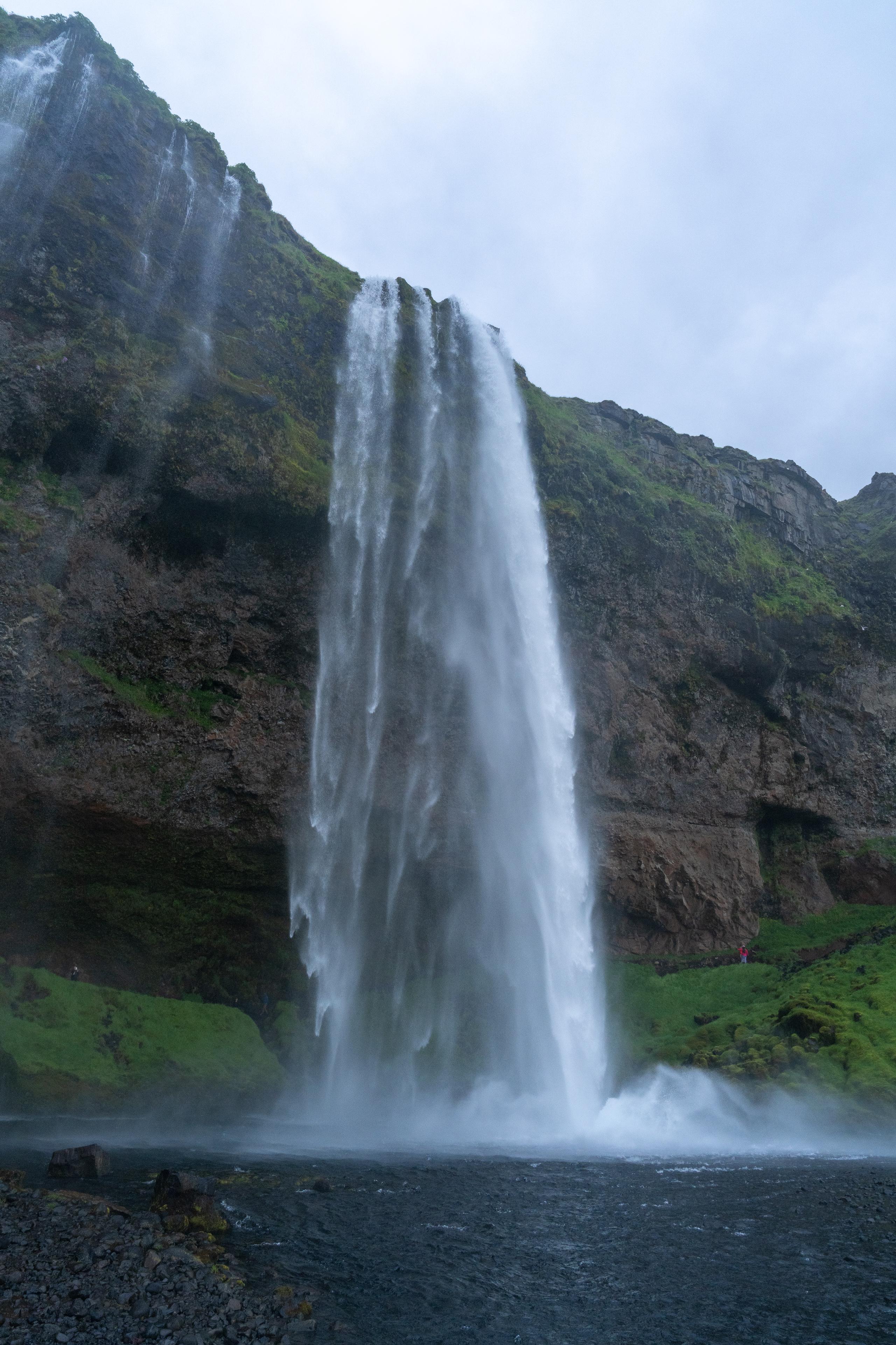 A tall waterfall falls vertically from a towering cliff. The water flows in a wide white water curtain, with large amounts of water mist and waves aroused at the bottom. The rock walls on both sides of the waterfall are dark brown basalt layers with green moss. A small green grass slope is exposed on the top of the cliff. The sky is gloomy gray-white. At the bottom of the frame are dark pools and gravel on the shore.