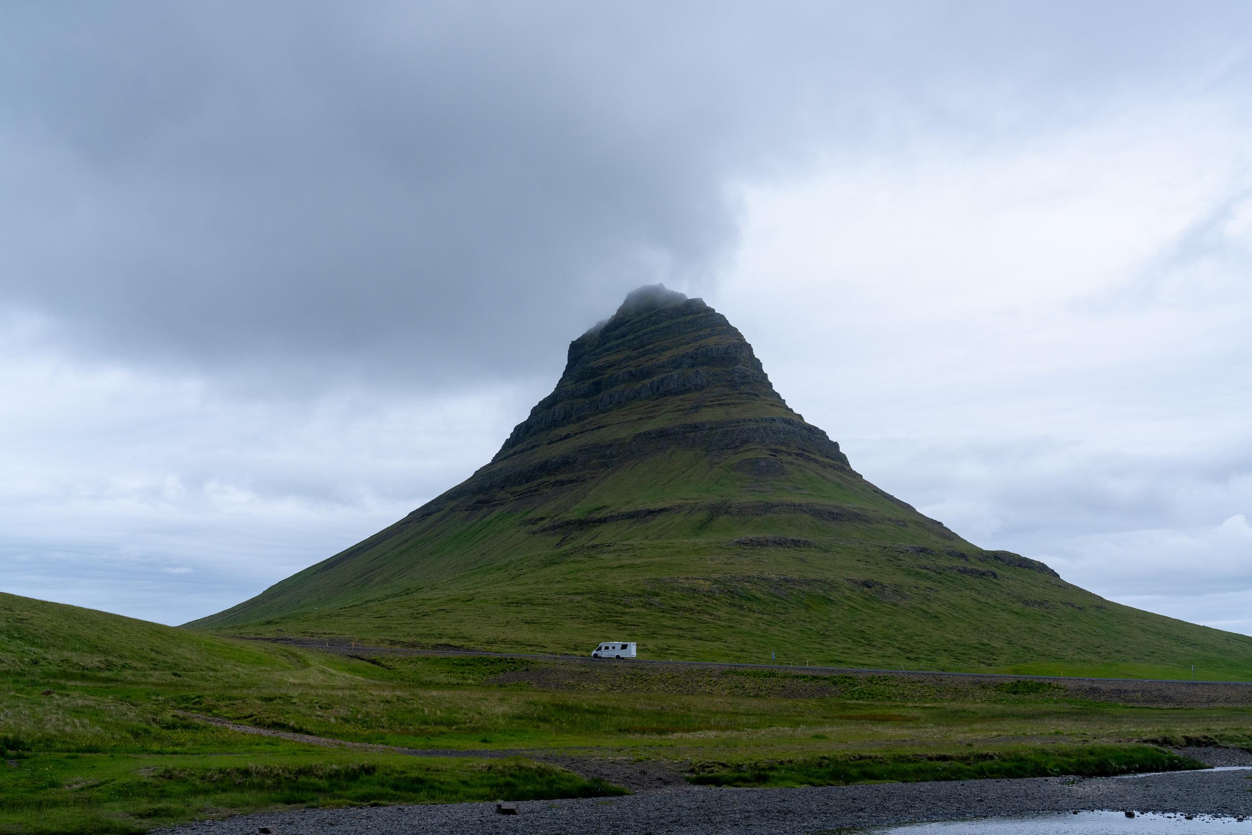 An isolated grassy mountain under a cloudy sky has a symmetrical cone shape. There are layers of horizontal rock formations and dark strips on the hillside, covered with green grass. The top of the mountain is covered by a low-hanging gray-white cloud. An asphalt road crosses the foot of the mountain, and a small white RV is driving on the road. The foreground of the frame is a green wetland, a creek and a gravel beach, and the sky is covered with thick gray clouds.