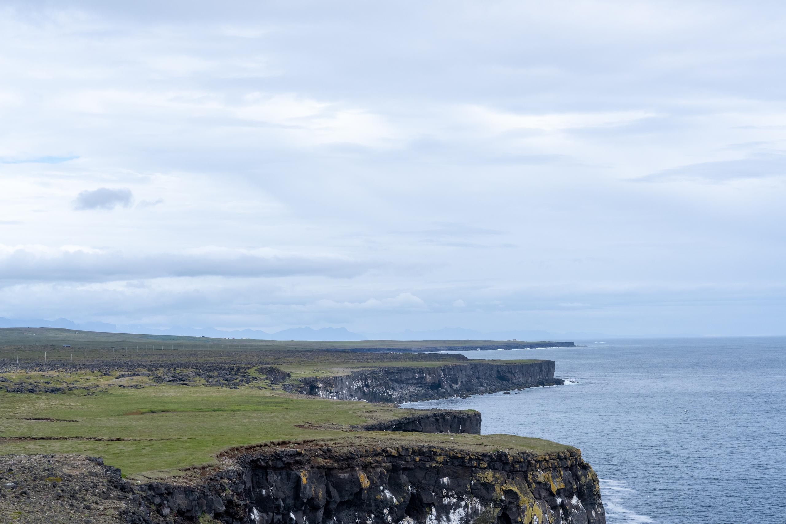 Broad coastline: In the middle of the frame is a long black rock cliff with a flat top. The top of the cliff is covered with green meadows and tundra. The cliff is dark and steep, with white waves lapping at the bottom. There is a vague road and telegraph poles on the flat field on the top of the cliff, and there are low mountain shadows on the horizon in the distance. On the right is the gray-blue sea, and the sky is full of clouds.