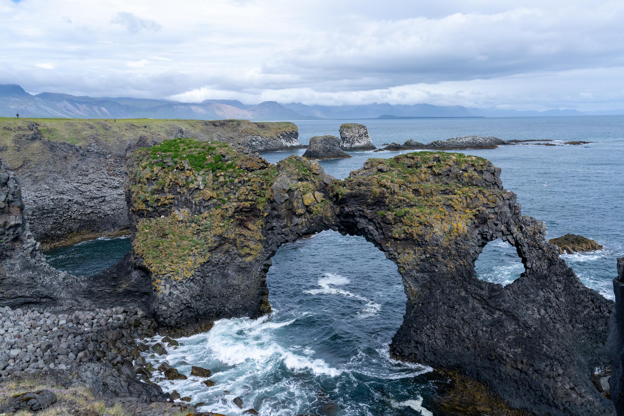 There is a natural arched cave on the huge black rock on the coast. The sea water passes under the cave and stirs up white waves. The top of the arched rock is covered with green grass and yellow-green moss, and the rock surface is dark volcanic rock full of holes. There are similar black rocks and grass slopes on the left and right sides of the frame. There are several small reefs scattered on the sea in the distance. There are continuous blue mountain shadows on the horizon, and the clouds in the sky are brighter.