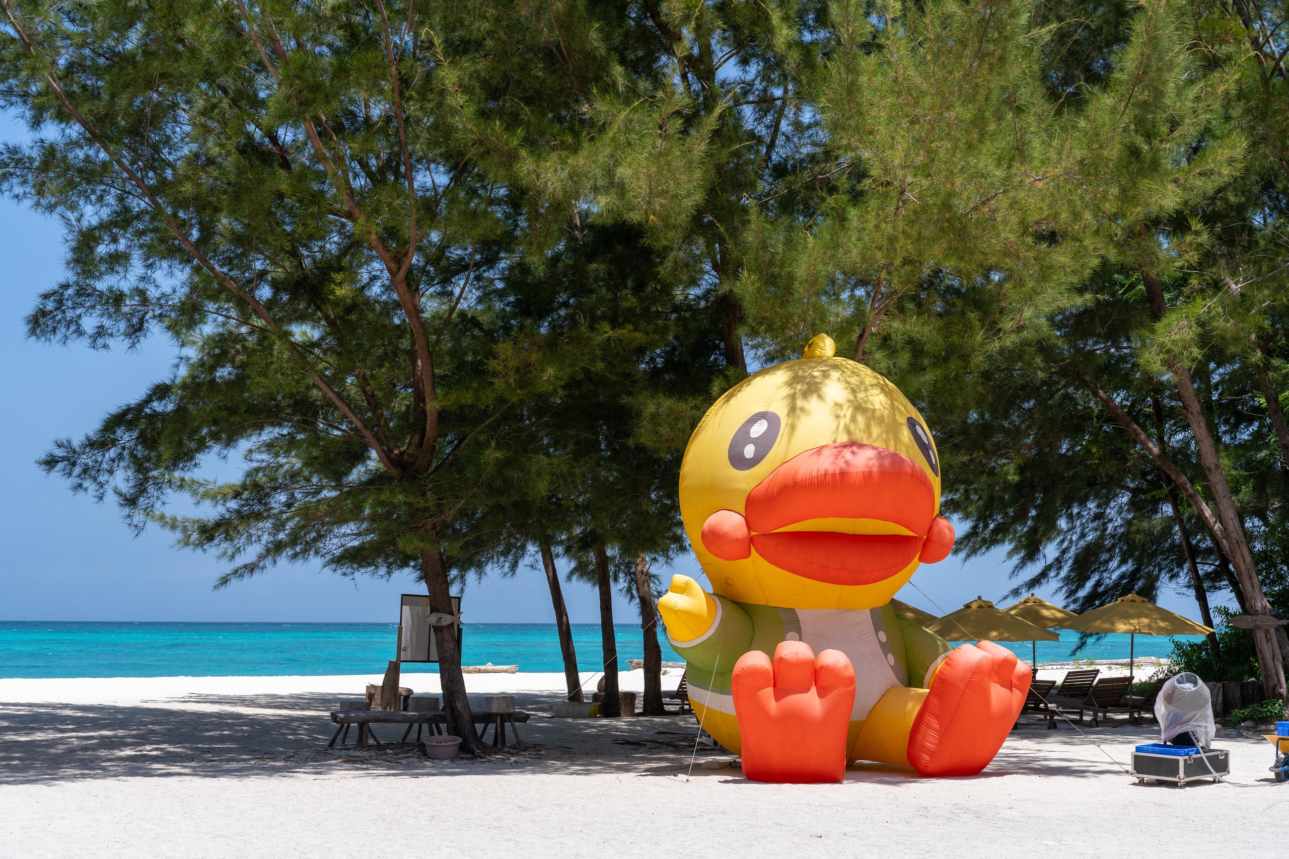 There is a huge inflatable yellow rubber duck propped on the beach, with an orange-red beak and soles, 'hands' on both sides of the body raised, and a white chest. Behind and on both sides of the duck are green sea pine trees with drooping leaves. There are parasols, deck chairs, and folding tables and chairs under the trees. You can see the turquoise sea through the tree trunks in the distance, and the beach is white.