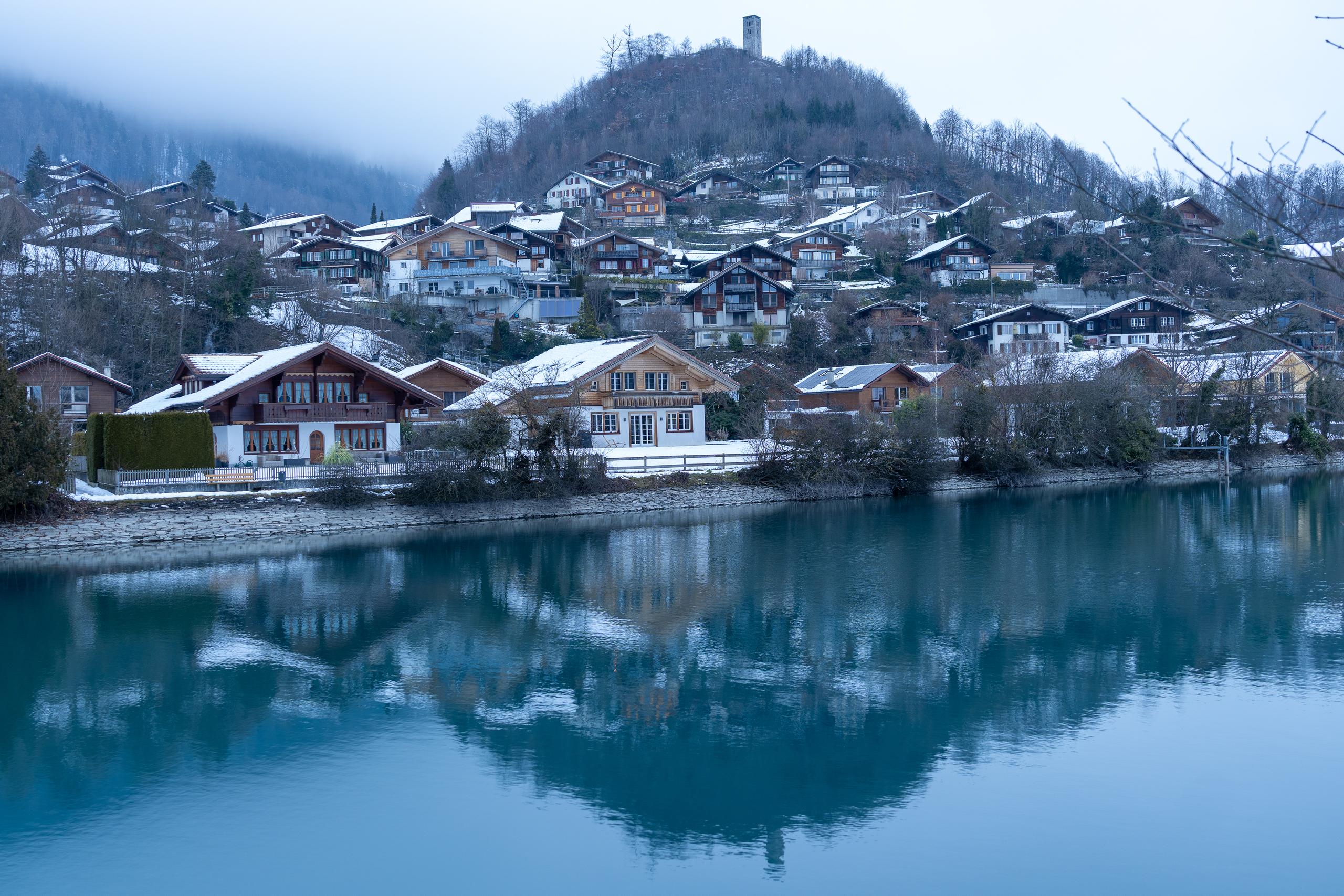 The lake is like a mirror, and the hillside on the other side is densely dotted with village houses with pitched roofs. The roofs are intertwined with fresh snow, and the woods and sky on the upper part of the hillside are shrouded in mist. The villages and woods are reflected in the dark blue lake below, almost completely symmetrical, and a small tower-like building stands on the top of the mountain.