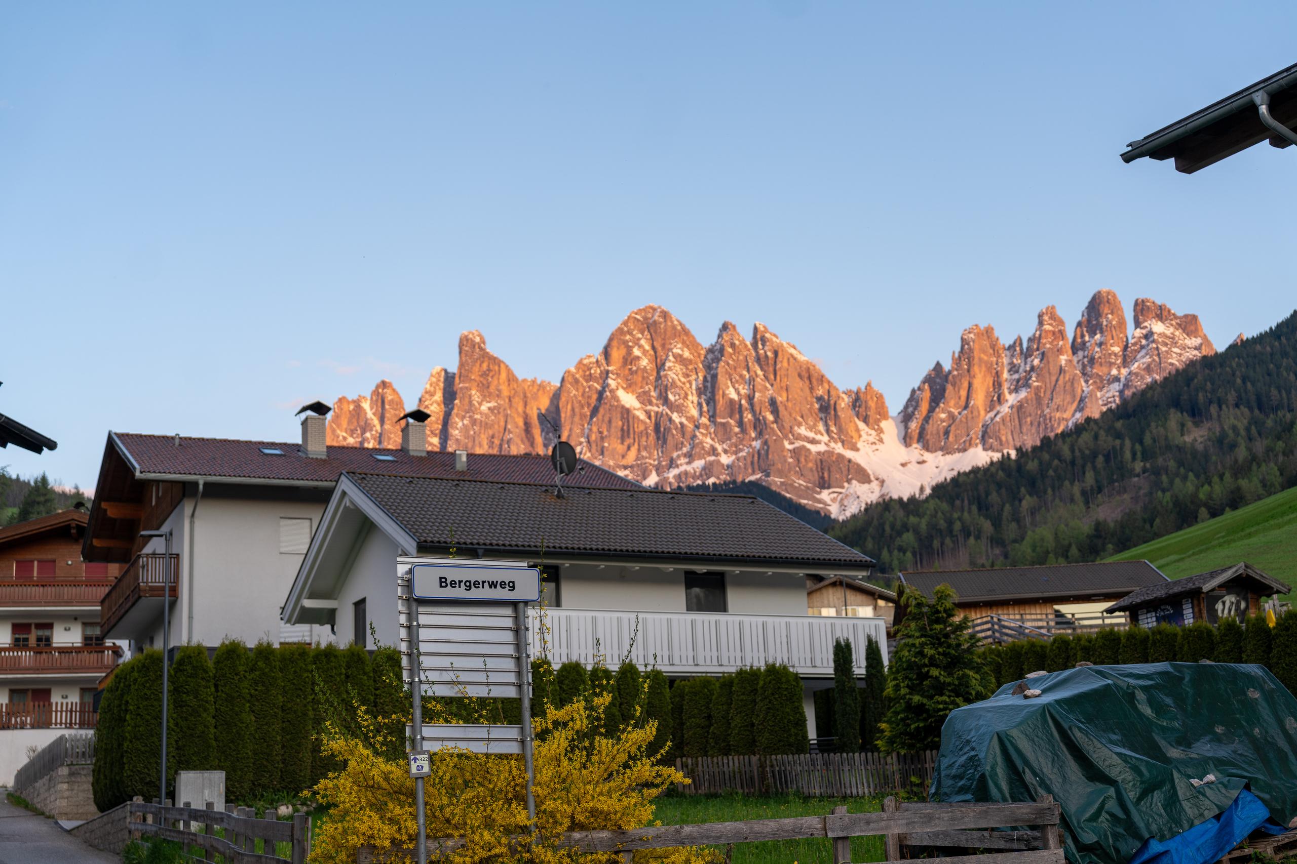 At dusk, a row of jagged rock peaks were dyed orange-red by the setting sun. There was still snow on the peaks, and the slopes below the peaks were covered with dark green coniferous forests. There was a village in the foreground, with two-story houses with white walls and deep roofs arranged at random. There were hedges and shrubs with yellow flowers in front of the houses. There was a street sign with the name of the street at the intersection, and a car on the right was covered by a green tarpaulin.