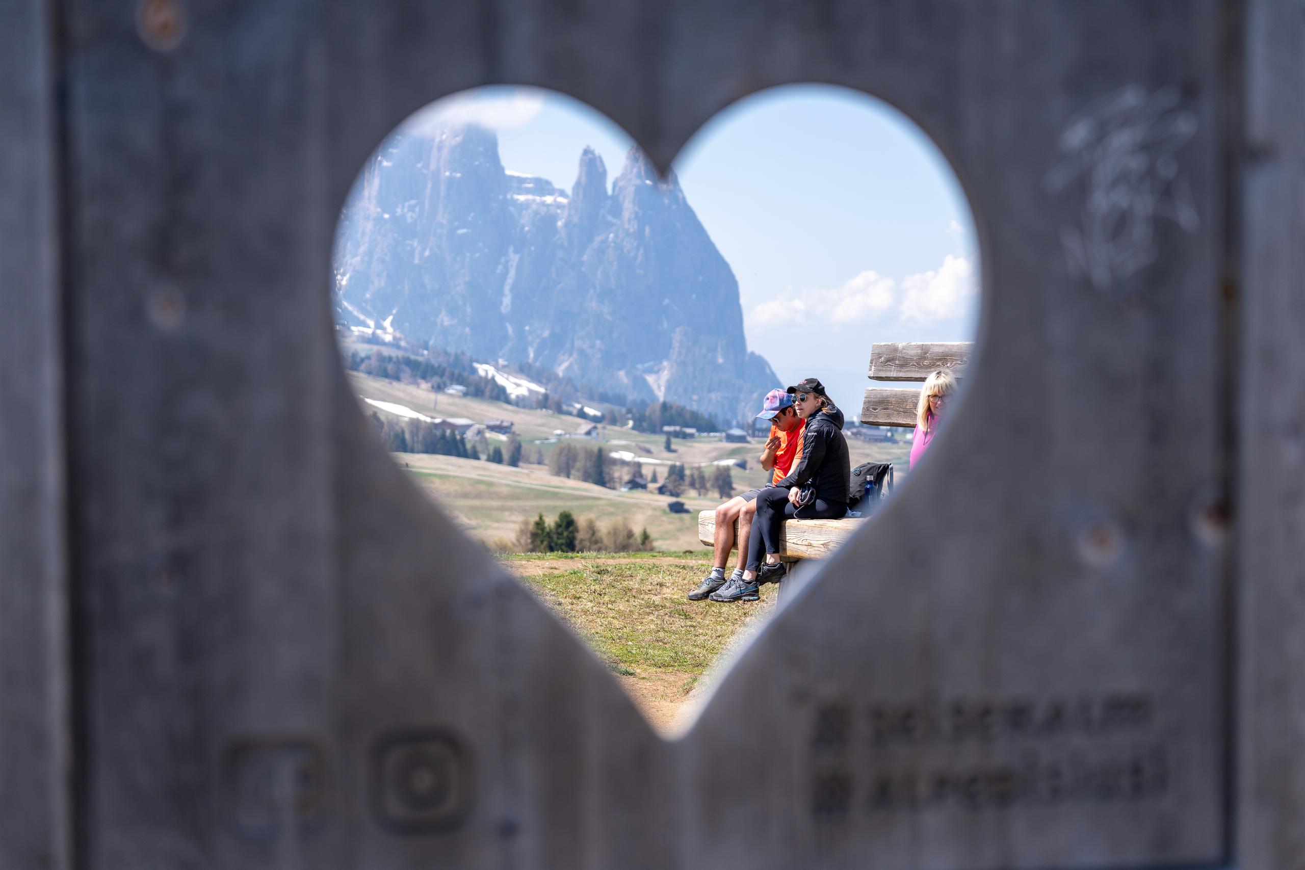 Looking through a heart-shaped hollow dug out of a wooden board, the middle scene shows a pair of men and women sitting on a wooden bench. One is wearing a black coat and a black hat, and the other is wearing an orange-red top. In the farthest distance is a huge light gray rock mountain peak; on the hillside are green and yellow meadows and sparse coniferous trees. Icons and lettering are faintly visible on the lower edge of the board.