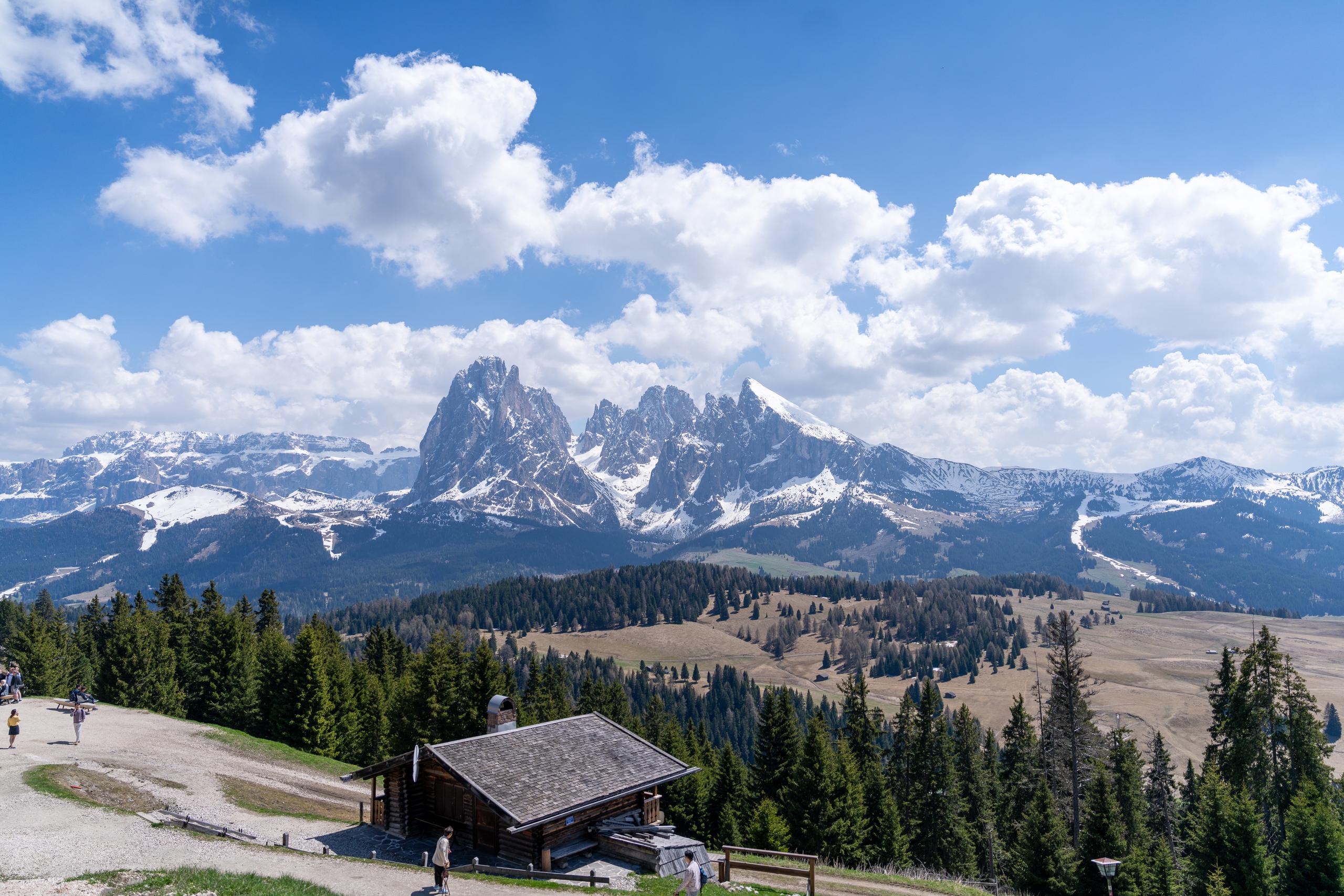 Panoramic view on the plateau: in the foreground is a dark wooden house with a gray wooden roof, and a few people walking on the gravel open space in front of the house; in the middle ground are rolling meadows and coniferous forests, in the background is a row of jagged gray-white rock peaks covered with residual snow, and the sky is blue and covered with large white clouds.