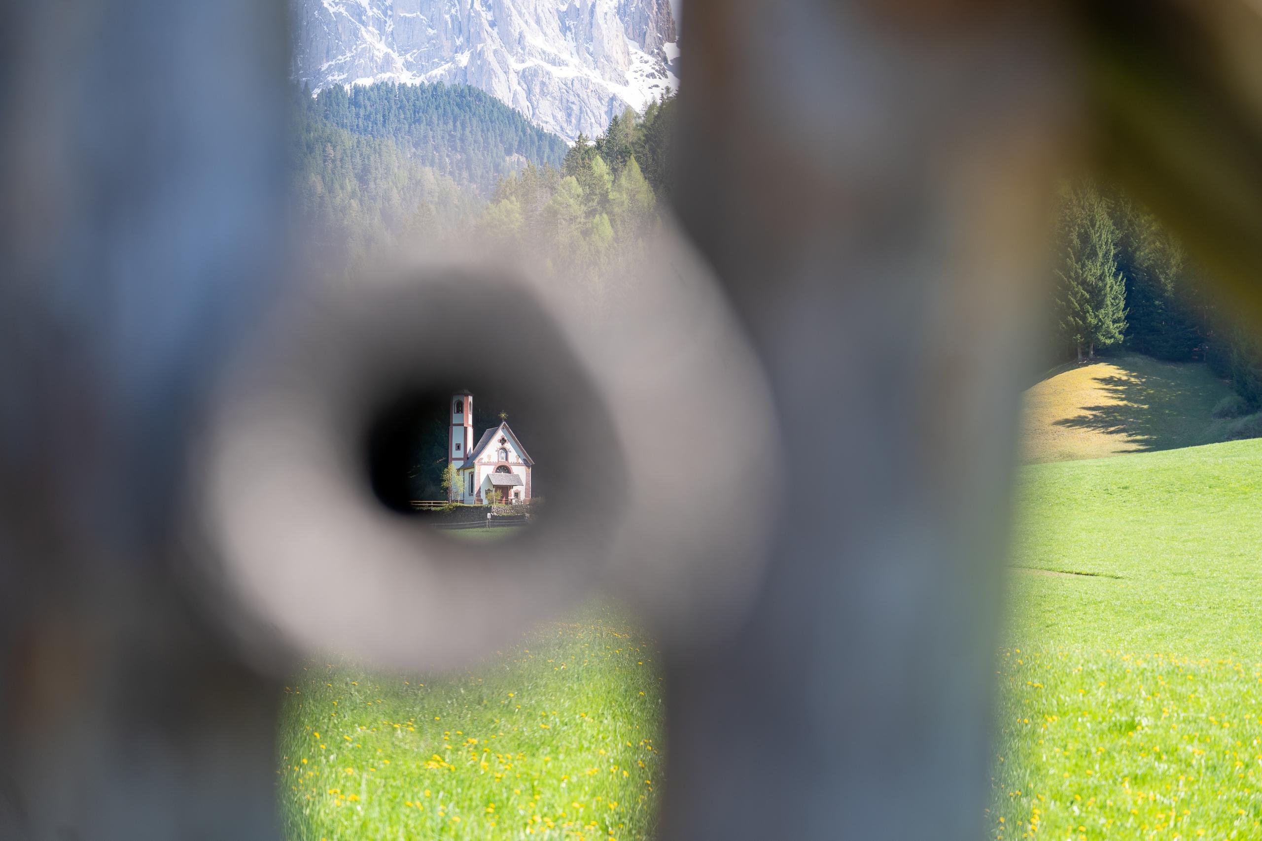 Looking into the distance through a strongly blurred circular hole: there is a small church with white walls and a red spire bell tower on the green grass in the center of the frame; behind the church is a dense coniferous forest and a steep light gray rock mountain peak, with small yellow flowers scattered on the grass.