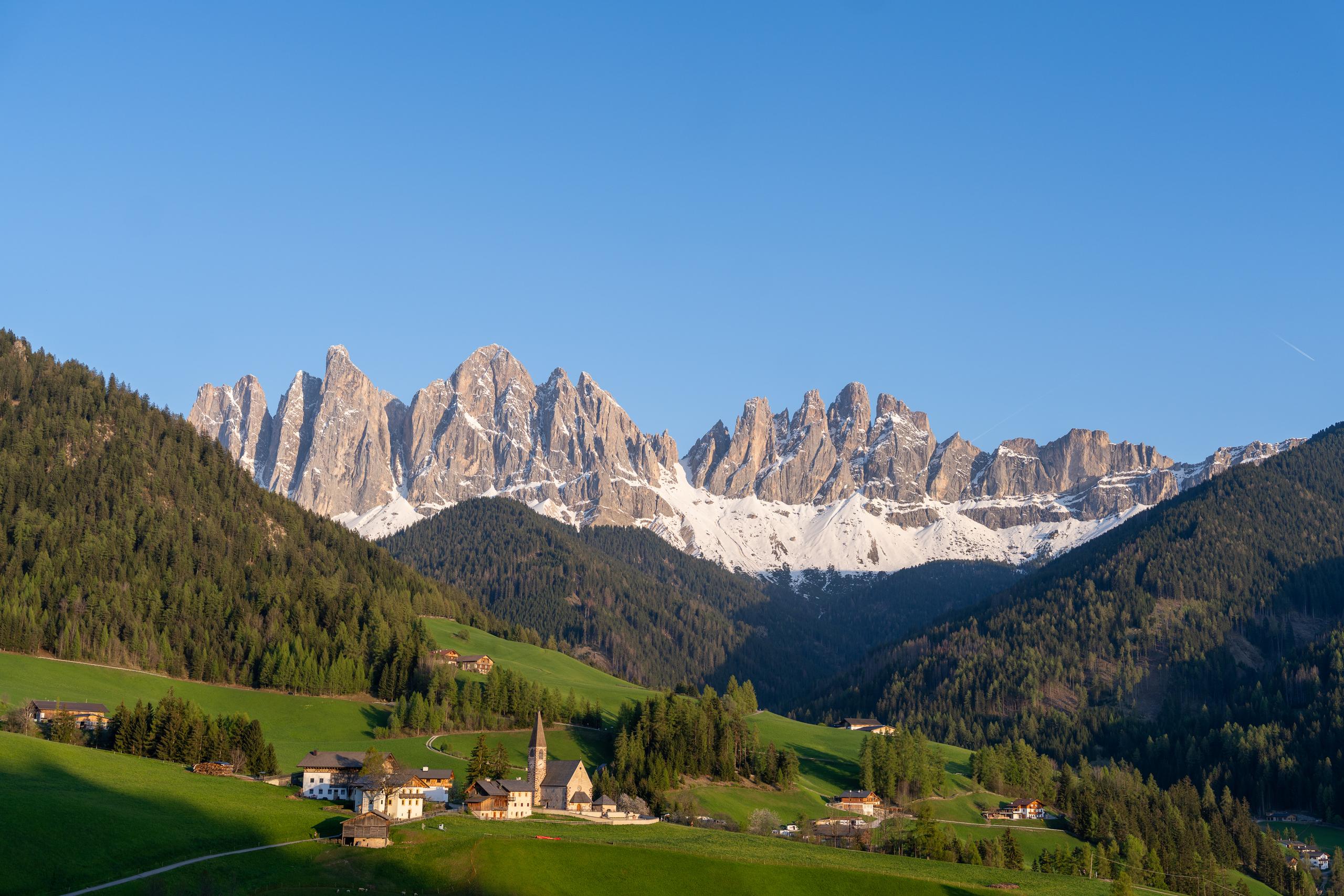 In the evening or early morning light, a large row of tooth-shaped gray-white rock peaks spans the upper part of the frame, with patches of snow hanging on the rock walls; in front of the peaks are rolling hillsides covered with dark green coniferous forests, and in the lower part of the frame is a bright alpine meadow. The meadow is dotted with cottages with white walls and deep roofs, and there is a church with a steeple in the center.