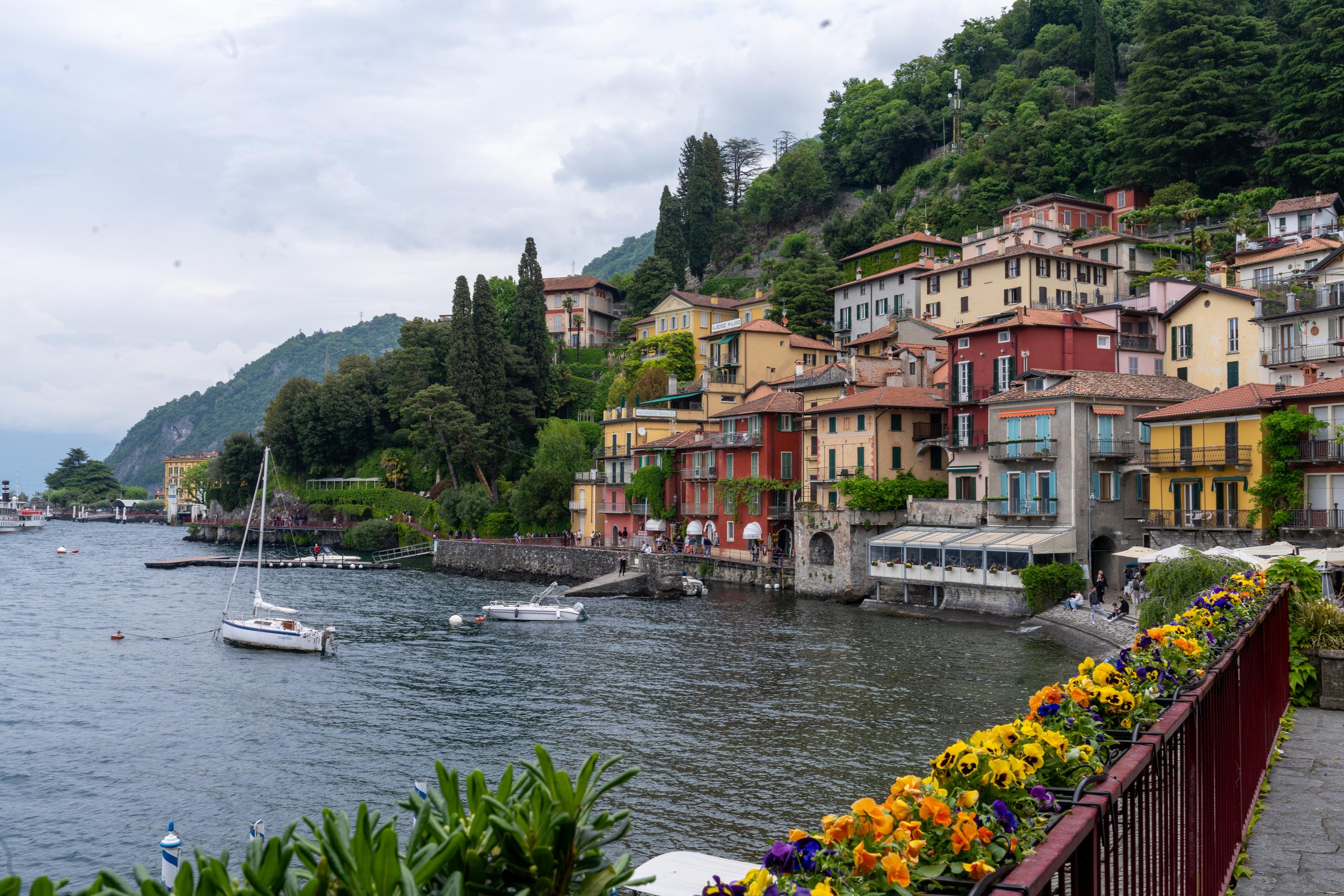 In a lakeside town under a cloudy sky, multi-story houses with yellow, orange, red and pink exteriors are densely stacked on a steep hillside, with green shutters on the windows; there are several small sailboats parked on the lake, and there are stone walls and arched waterways on the shore; in the lower right of the foreground are red railings and a long row of yellow-purple flowers in full bloom, and in the distance are mountain forests in the mist.