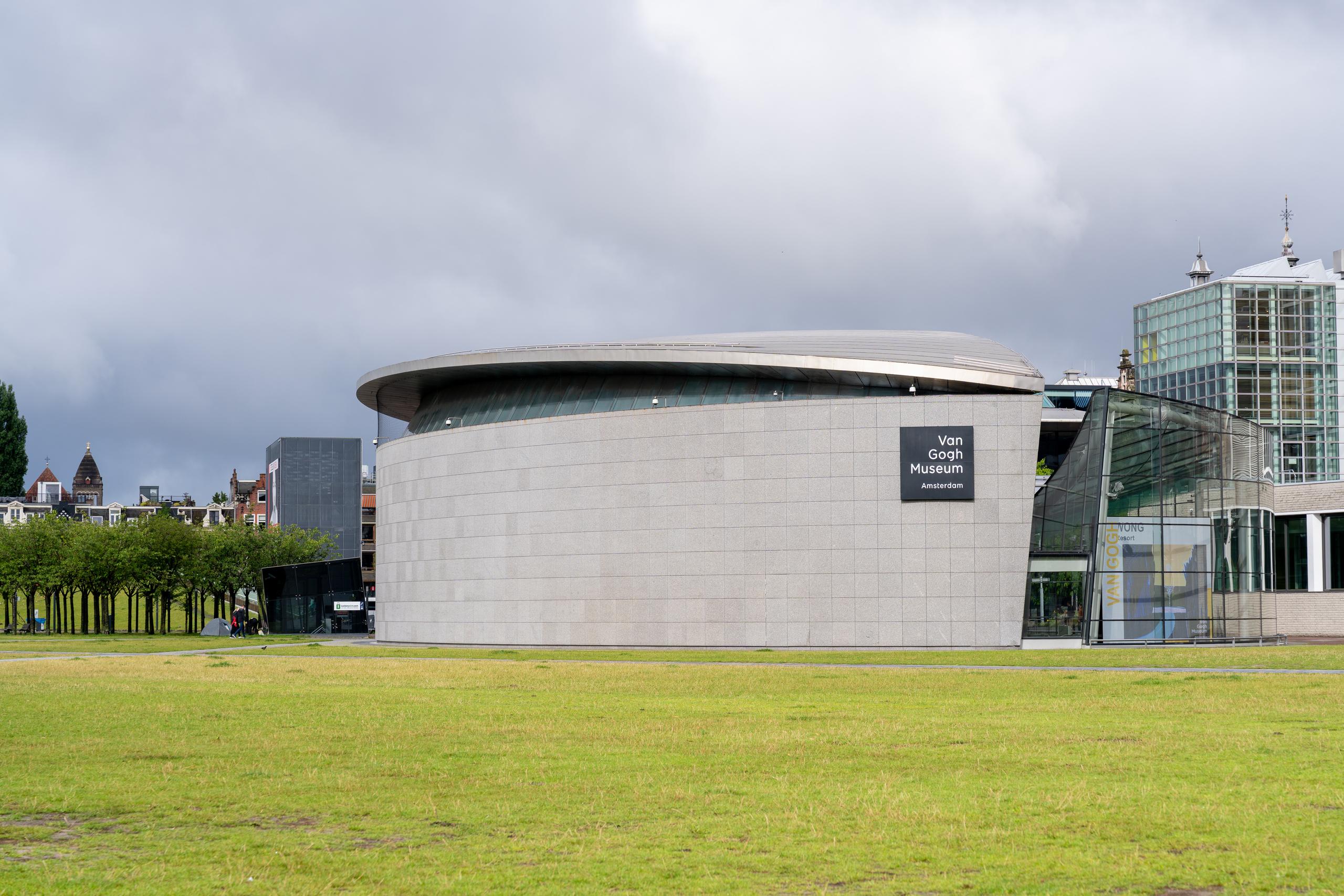 The appearance of a modern art museum under a cloudy sky: the main body is a curved building with a translucent light-colored stone exterior wall and a dark oval eaves on the top. A signboard with the name of the museum in white letters on a black background is hung on the curved wall; on the right side of the building is a connected glass curtain wall part, and the internal stairs can be seen; on the left background are red old-fashioned spire houses and rows of green trees, and the sky is covered with dark clouds.