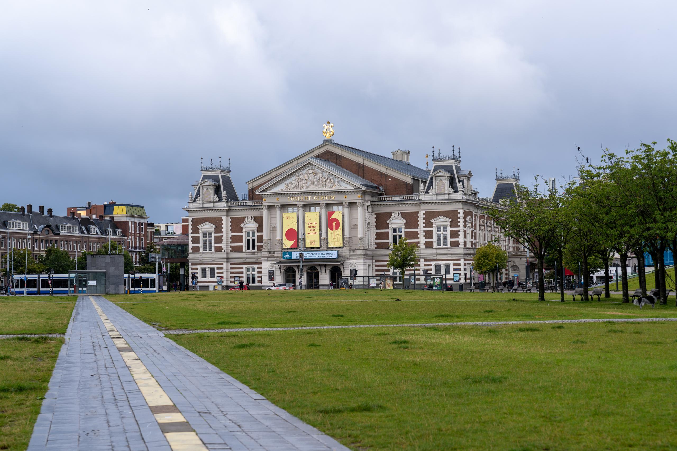 The square and classical buildings on a cloudy day: in the central background is a majestic brick-red classical theater, with a pediment and rows of sculptural reliefs on the front, golden decorations on the edge of the roof, and large colorful performance posters hanging on the walls; in front of the building are broad lawns and trees, with a blue-and-white painted tram on the left; a paved brick walkway extends toward the building in the lower left corner.