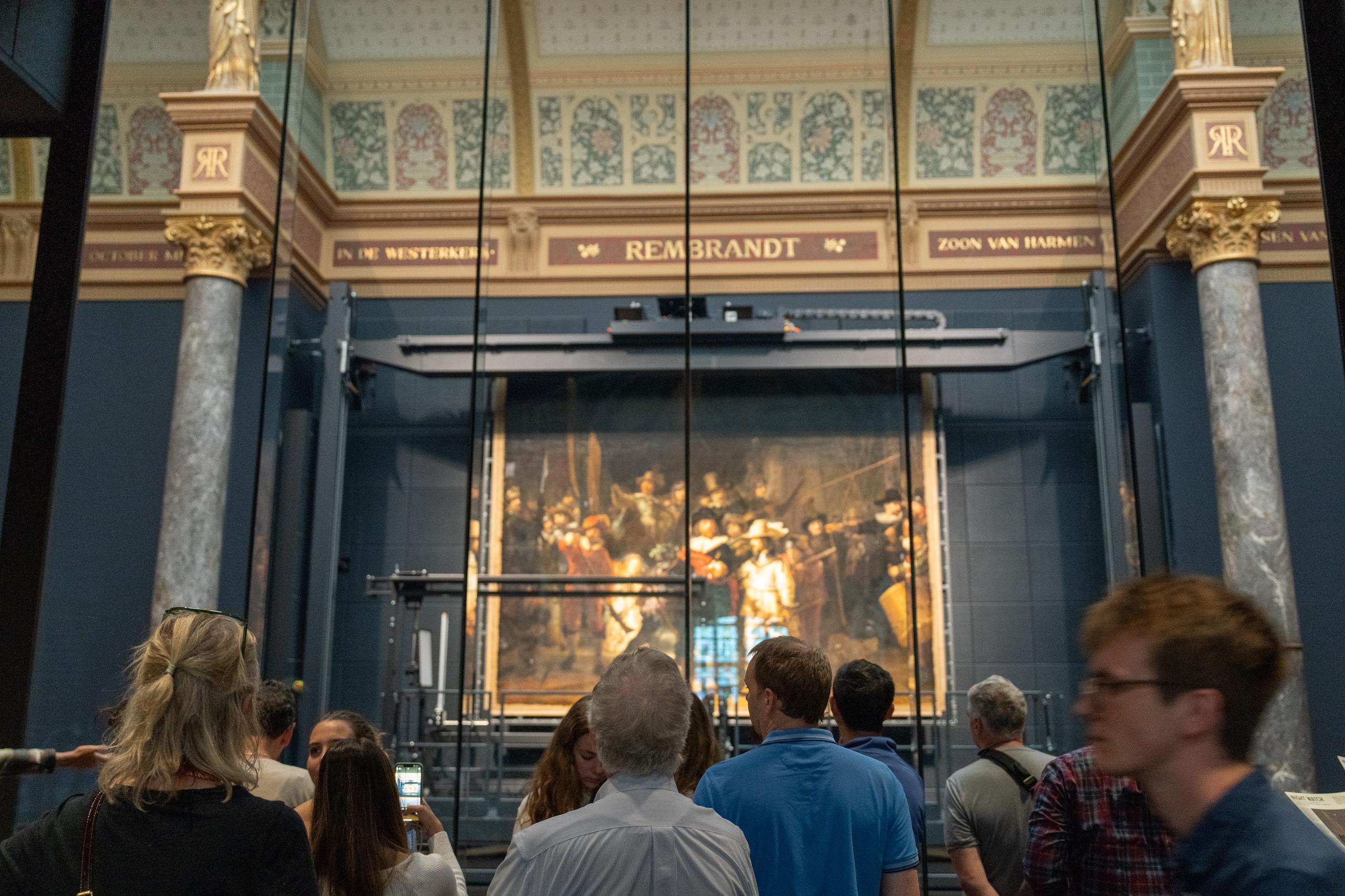 There is a high-open exhibition hall in the art museum: a huge classic group portrait oil painting hangs on the wall in front, with many characters and a dark and golden tone. The painting is mounted behind a glass partition wall. Through the glass, you can see the reflection of the bracket in front of the painting and the audience; there is a banner above the wall with the name of the artist; in the foreground is a group of spectators with their backs to the camera, one of whom is holding up a mobile phone to take pictures.
