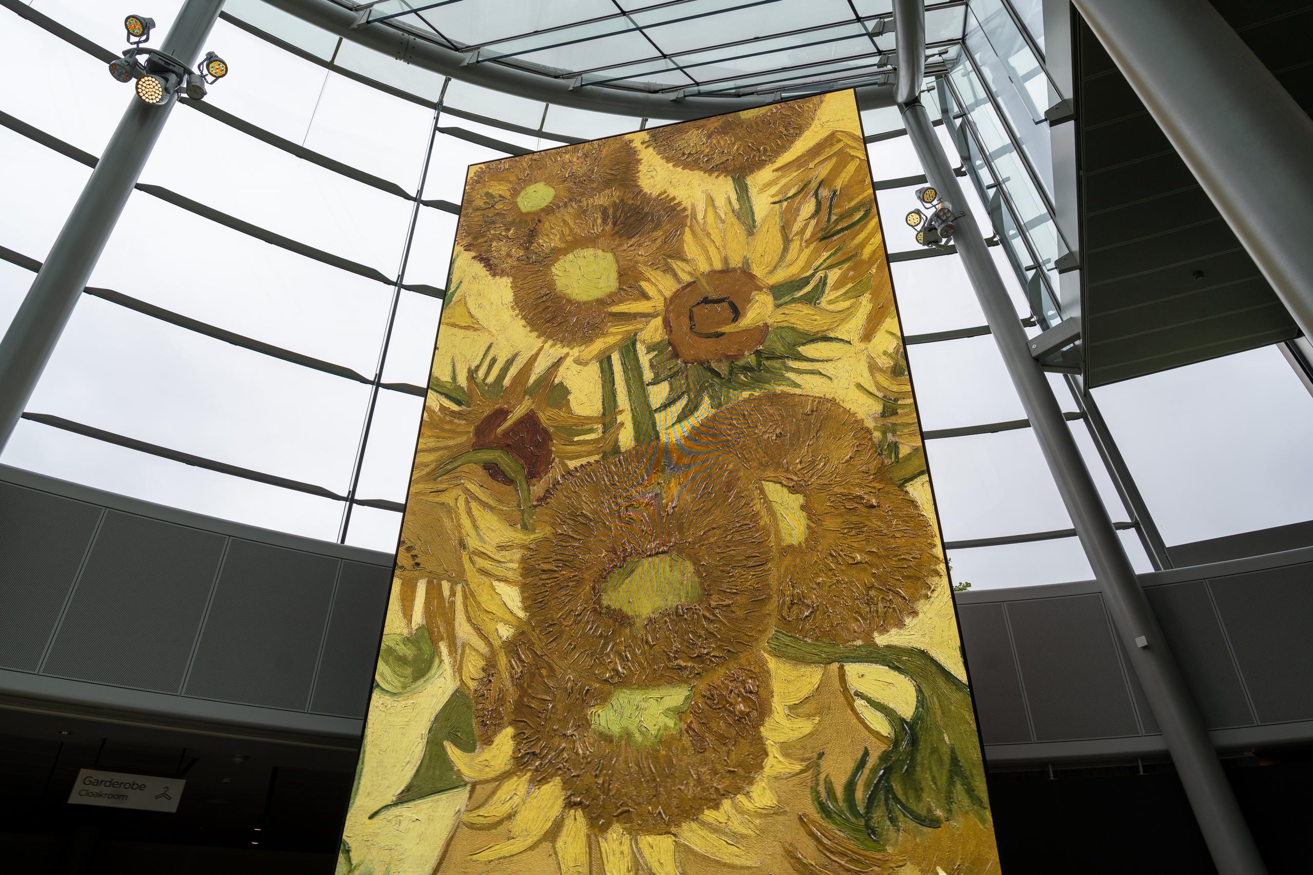 Looking up from a low angle at a large painting hanging in a tall glass curtain wall: in the center of the painting is a long vertical yellow-toned painting of a sunflower in bloom, with flowers and leaves expressed with thick brushstrokes; behind the painting is a curved glass curtain wall, through which the gray sky and metal frames can be seen, and several spotlights are mounted above the painting.