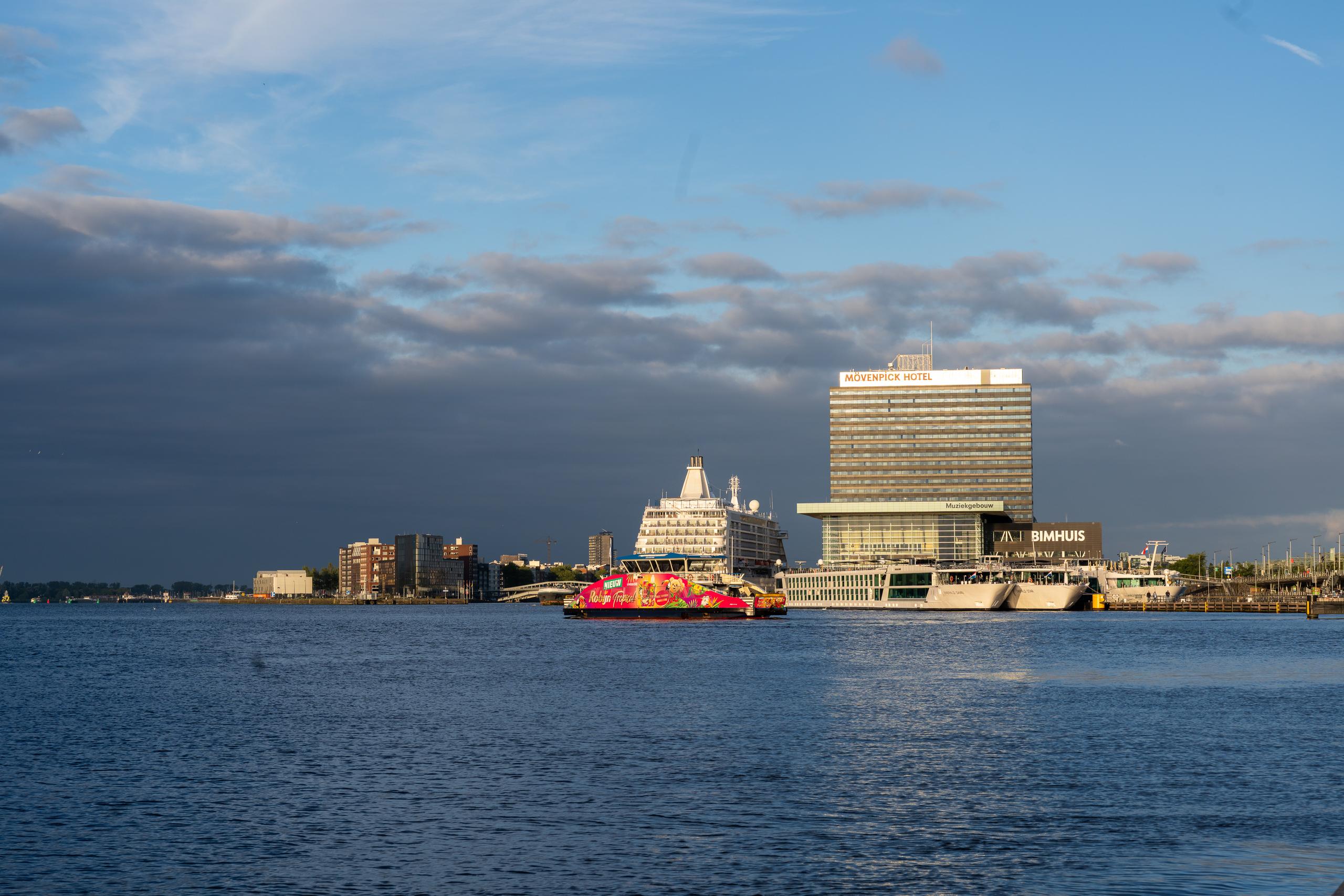 The water and the city pier in the evening: On the left side of the center of the frame, there is a bright pink round-headed small cruise ship parked on the water, with decorative paintings on the side; on the right is the pier area, with a high-rise modern board-type hotel building, in front of the building are low glass buildings and dark buildings with words; in the distance on the left are low shorelines and cranes, on the left side of the sky are thick dark gray rain clouds, and on the right side is the blue sky.