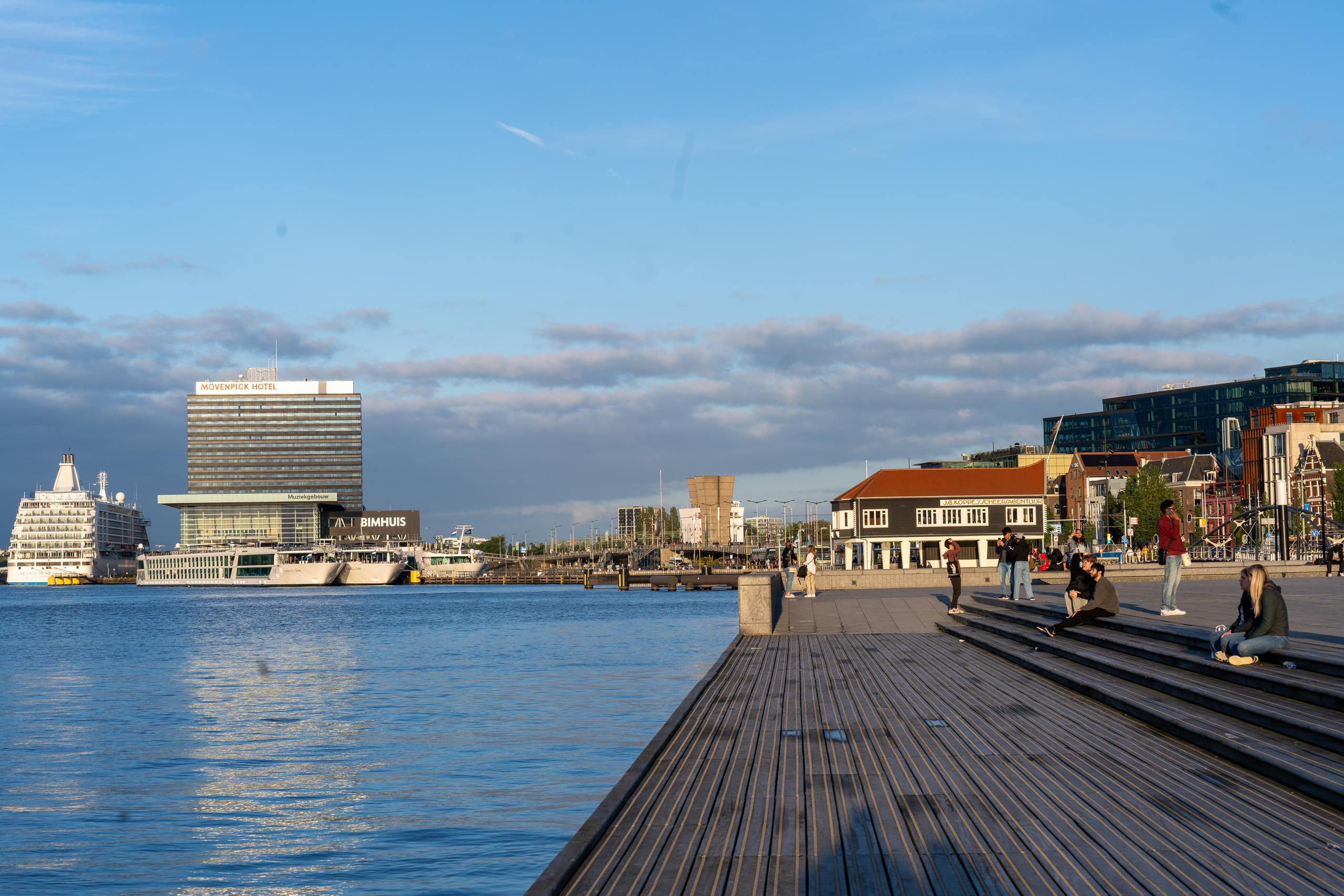 City waterfront in the evening: On the left is the open water, rippled and reflecting the sky; on the opposite bank is a high-rise modern panel hotel building and the low glass building in front of it, and a white multi-story cruise ship parked further to the left; on the right and lower right is a wide water-friendly platform paved with dark wooden strips, where several people sit on the wooden steps to rest and chat, and in the distance there is a red-brown sloping roof building.