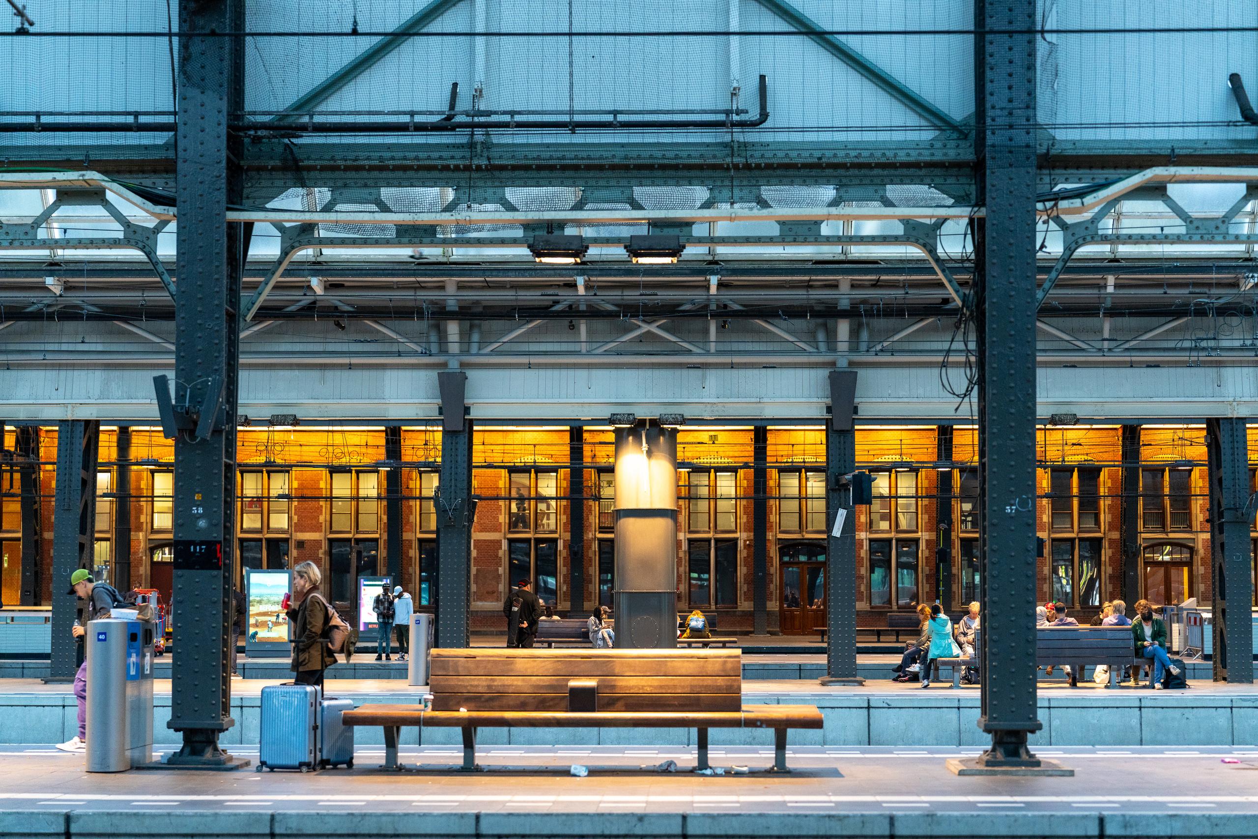 The platform of an old-fashioned railway station: above is a dark green steel frame and glass canopy, with thick riveted steel columns standing on the platform; in the mid-ground is a row of wooden benches and a metal bus shelter. There are several trolley suitcases on the platform, and a long-haired lady is standing on the left pulling the suitcase; in the background is the brick station facade, with a row of arched doors and windows showing warm yellow light, and passengers waiting on the platform are scattered around.