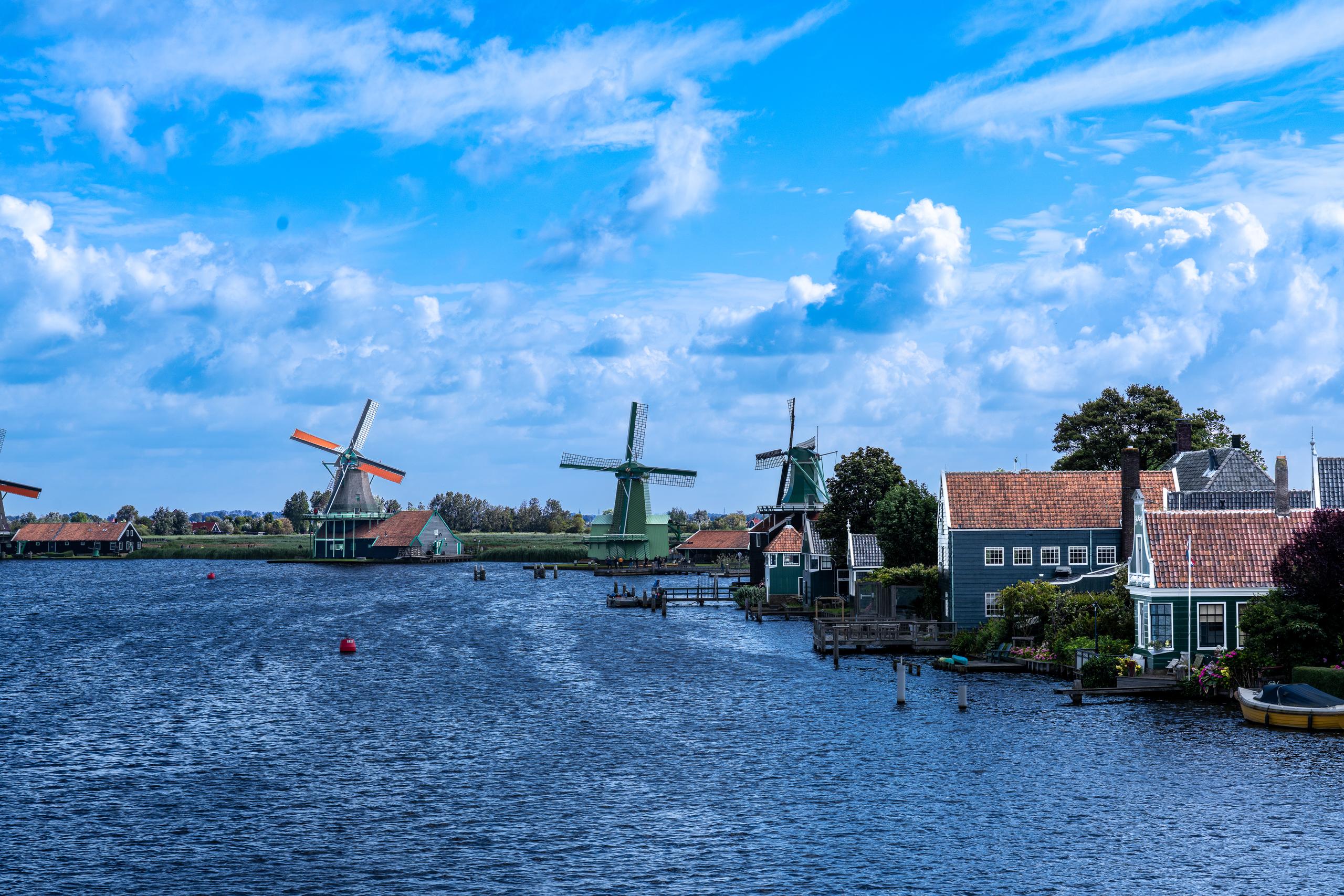 Canal and Windmill Village on a Sunny Day: In the middle of the frame is a wide canal, with dark wooden walls, green and white traditional wooden houses with red tile roofs on both sides; three tall traditional windmills stand on the bank, with white and green towers equipped with four long blades; in the distance, the low fields extend to the horizon, and the sky is blue with large cumulus clouds.