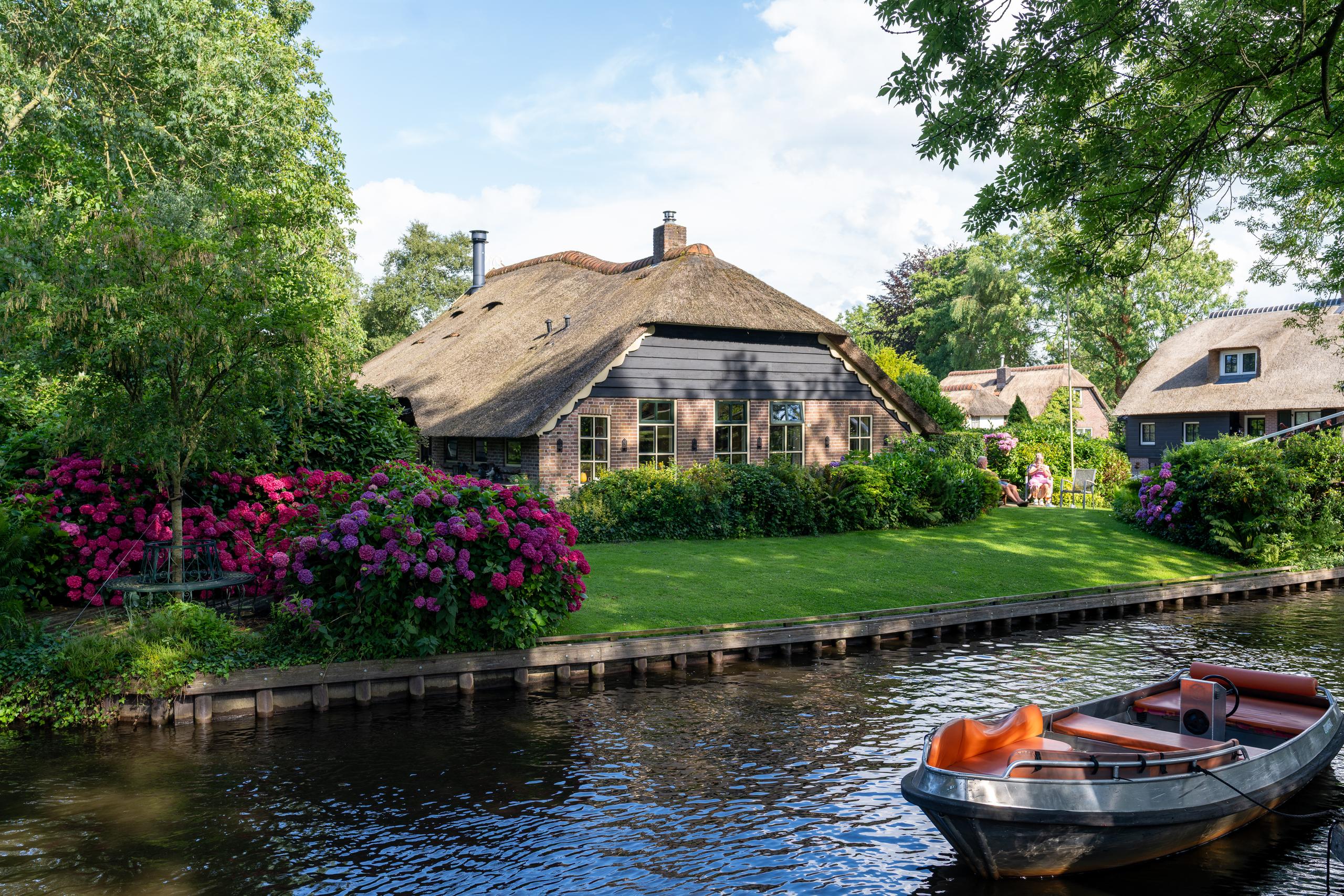 Houses on the canal under the sun: On the opposite bank is a traditional house with brick walls and a huge reed thatched roof. In front of the house is a neatly trimmed green lawn. On the left side of the lawn is a large cluster of pink and purple hydrangeas in bloom; the canal bank is reinforced with wooden piles. A wooden flat-bottomed boat is parked on the water in the lower right corner. There are orange-red seats and wooden backrests in the boat.