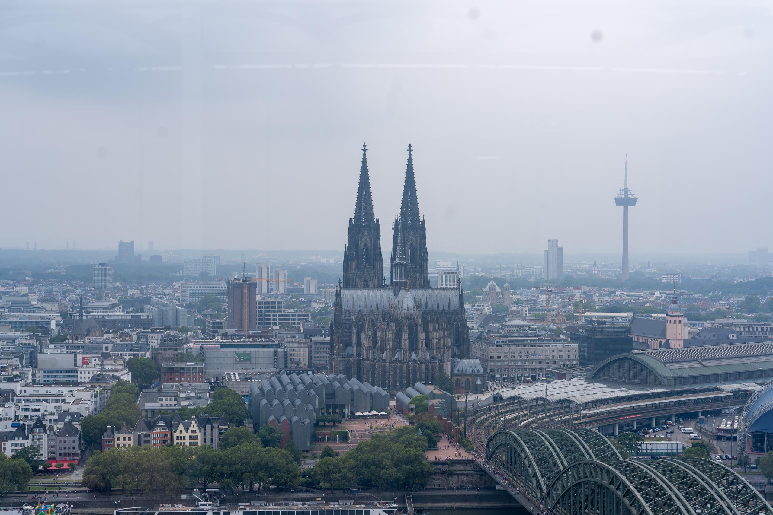 A panoramic view of the city from a high place on a cloudy day: in the center of the frame is a huge Gothic cathedral with two dark towering spiers reaching into the sky. The main body of the church is made of dark gray stone. It is surrounded by dense urban buildings and streets. A towering TV tower stands on the right. On the lower right is the curved glass roof of a large train station and an iron bridge across the river. The sky is gray and white.