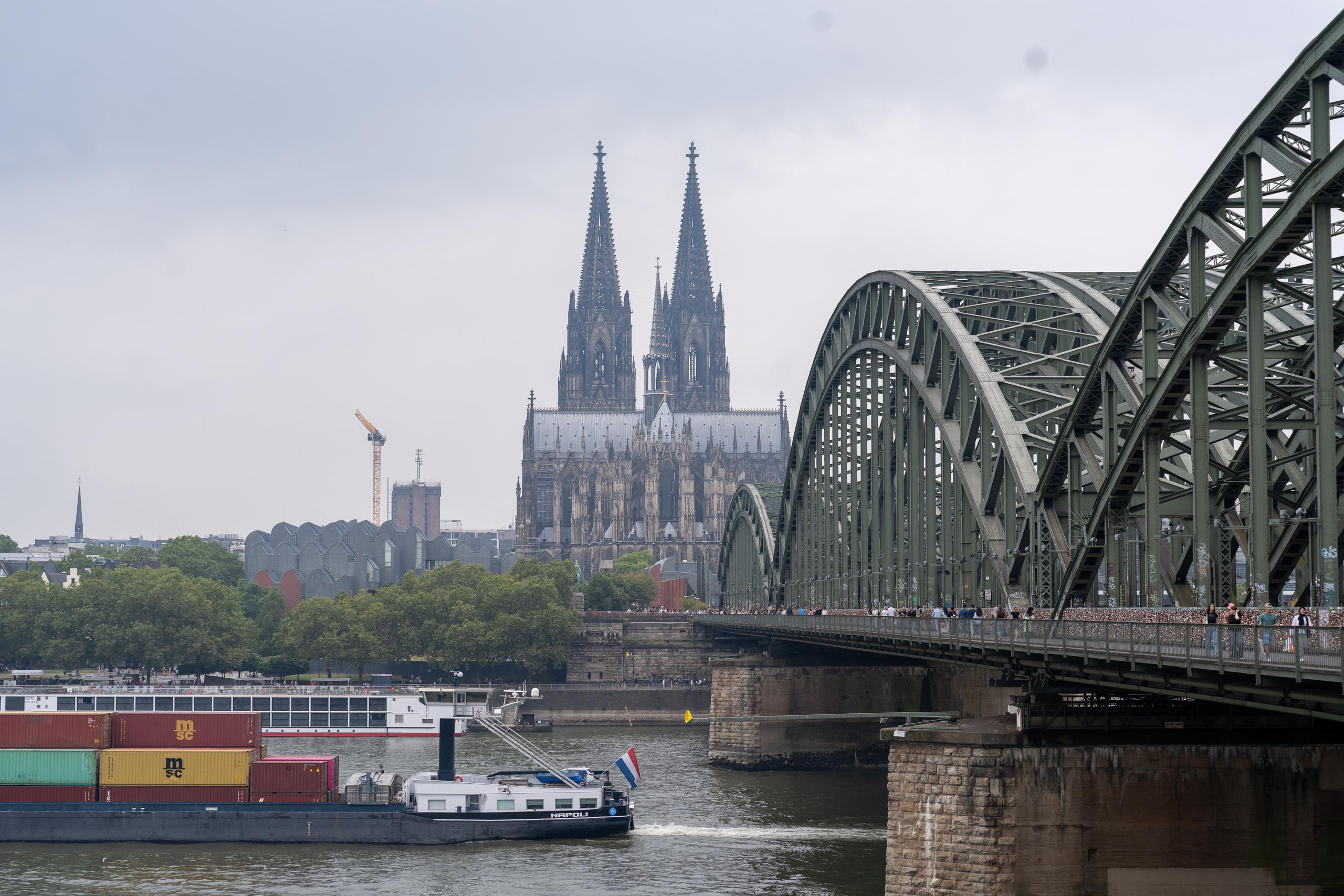 Riverside landscape on a cloudy day: The right half of the frame is a porous arch bridge with a green steel structure. The bridge body is composed of multiple arch trusses. There are many pedestrians walking on the bridge. The bridge piers are heavy stone structures. In the center of the background is a Gothic twin-tower cathedral, surrounded by green trees and urban buildings. A barge loaded with colorful containers is sailing on the river on the left.