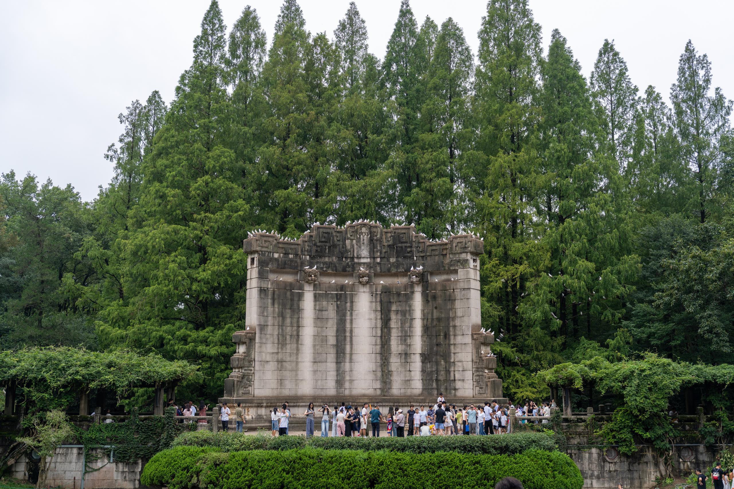 A large light gray stone monumental building is located in front of a green forest: the monument is rectangular, with a crest on the top and rows of carved decorations, and several vertical grooves on the surface; there are many white birds on the top and sides of the monument; there are dozens of tourists in summer clothes gathered on the platform in front of the monument, and some are taking a group photo; below the frame are neatly trimmed hedges and stone walls.
