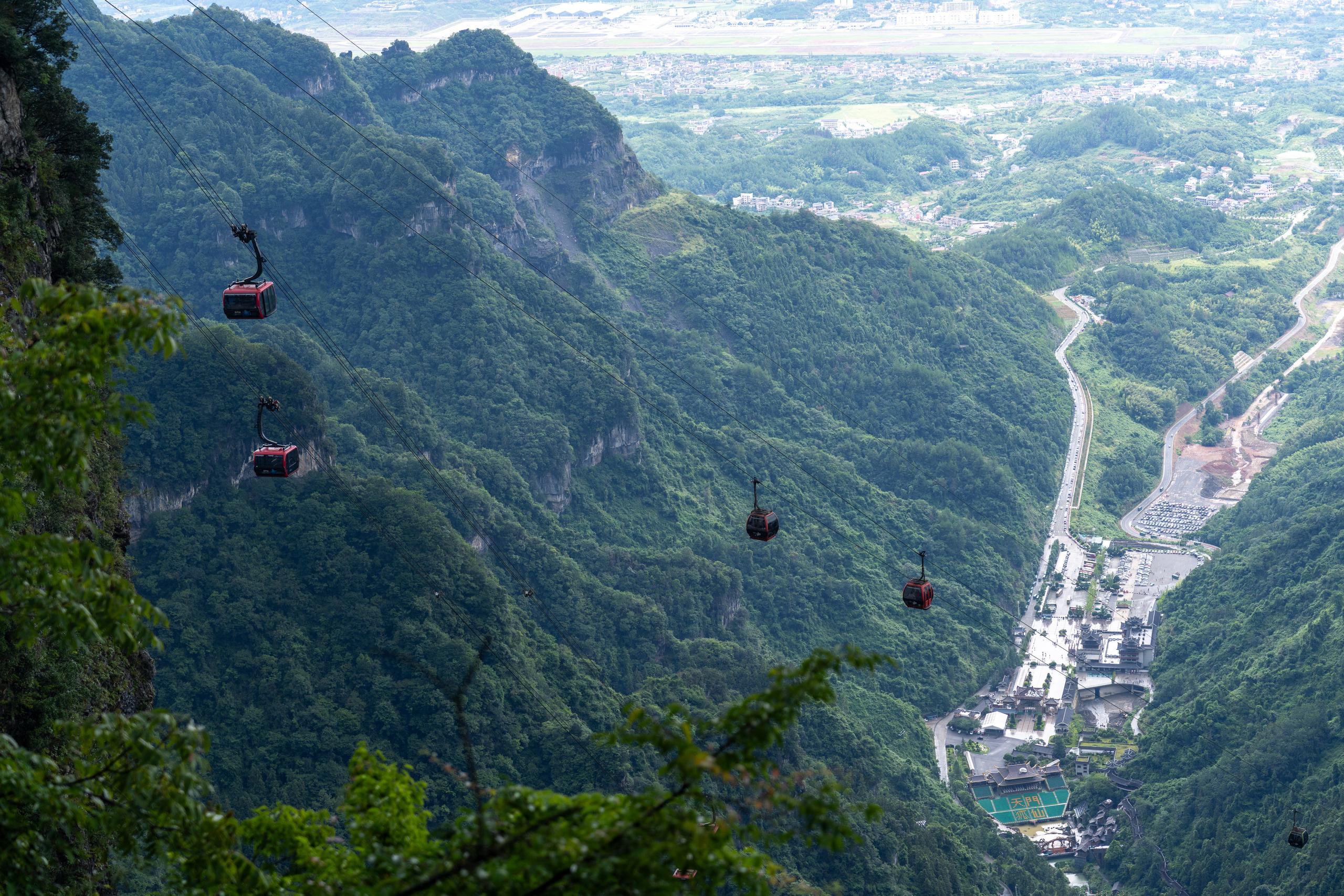A high-angle view of a mountainous canyon: several red cable cars are suspended on cableways, traversing high altitude along one side of the canyon, arranged diagonally in the frame; below are steep ridges and valleys covered with dense green forests, on the lower right is a winding road, a large parking lot and villages at the foot of the mountain, and in the distance are open plains and towns.