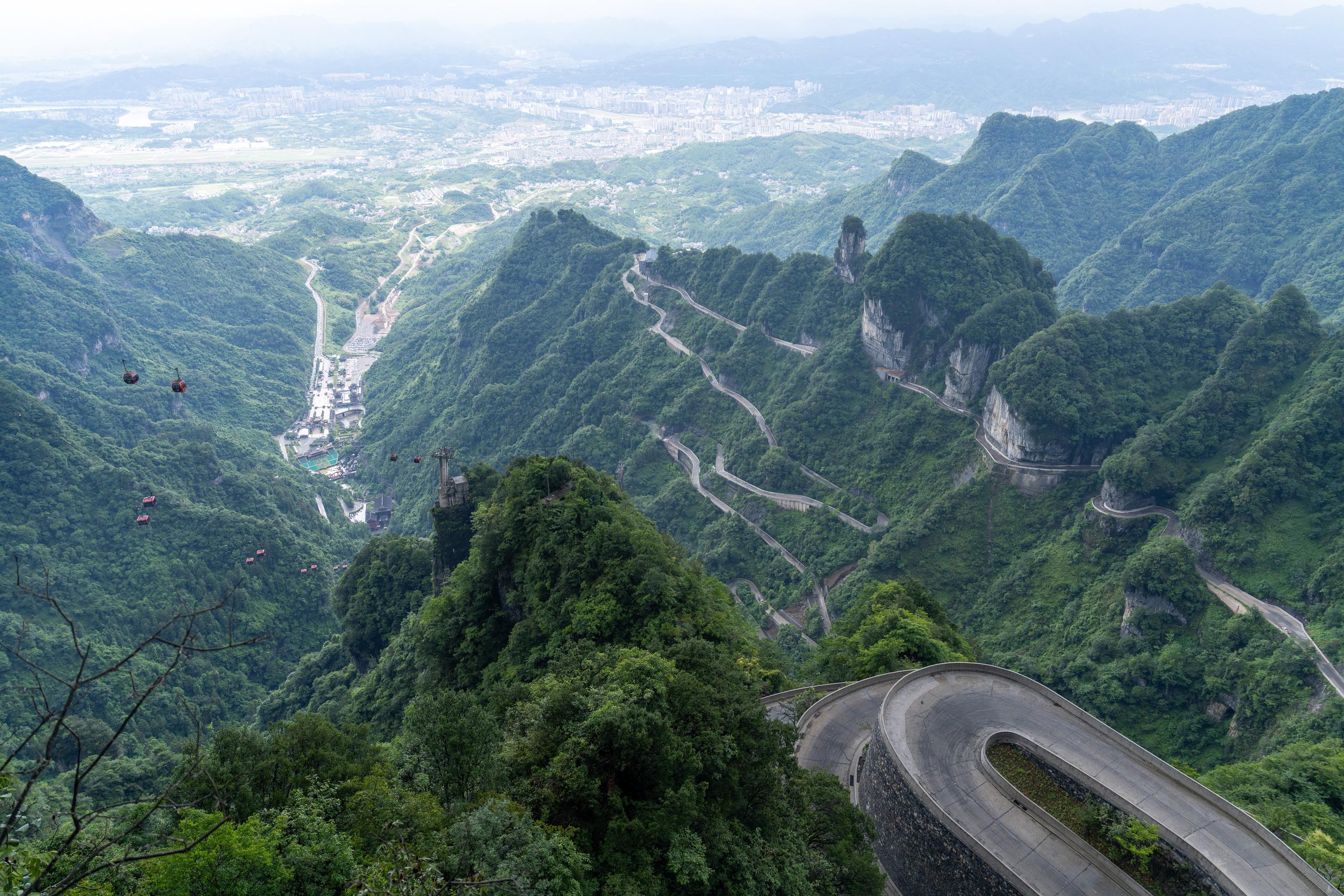 A panoramic view of the mountainous area from the top of the mountain: the frame is dotted with pinnacles covered by dark green vegetation, and a winding mountain road winds down the mountain with dense switchbacks; rows of cable car cableways and a group of buildings in the valley can be seen in the remote position on the lower left, and in the distance are cities and foggy outlines on the plain; the tops of several peaks in the close view are exposed rock pillars.