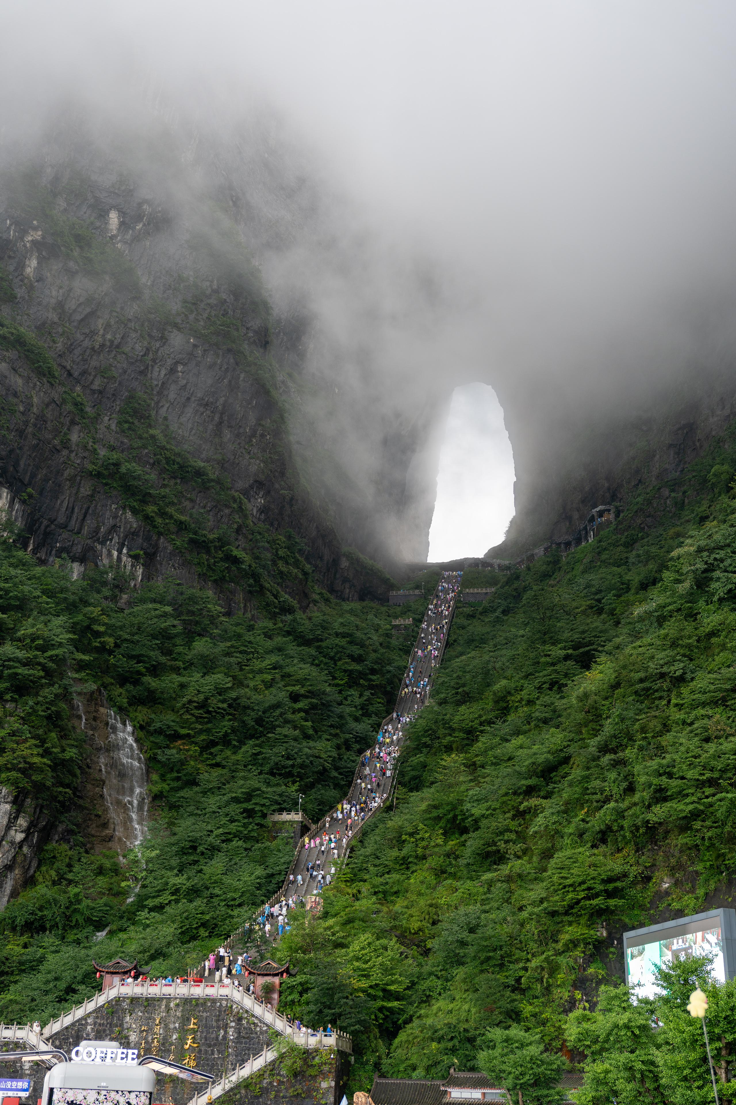 A natural mountain cave landscape: there is a large oval hole in the center of the huge mountain, with a bright white sky revealing the entrance, surrounded by dense white clouds; there is a steep and extremely long stone staircase from the foot of the mountain leading directly to the entrance of the cave, and the steps are densely packed with tourists climbing up; both sides of the mountain are covered with dense green vegetation, and a thin white waterfall flows down the left side.