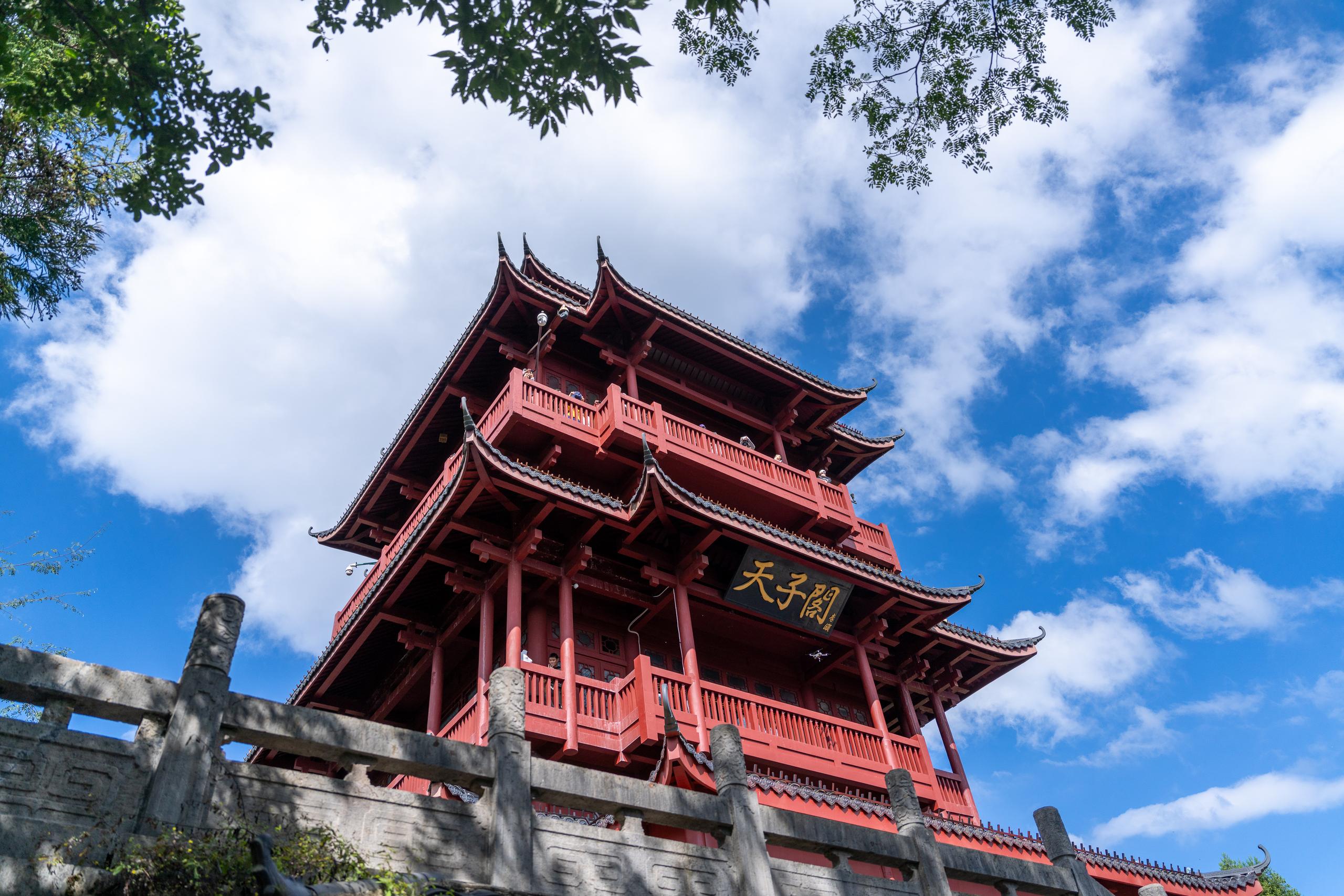 Looking up at a multi-story Chinese-style wooden pavilion: the pavilion is mainly bright red, with multiple overlapping eaves that retract upwards, and the ends of the eaves are raised. There are red railings and columns on each floor, and the bracket structure can be seen under the eaves; a dark blue background with gold letters hangs on the front of the pavilion; there is a gray-white stone guardrail on the left side of the foreground, and leaves extend into the upper right corner of the frame; the sky is blue, with large white clouds floating.