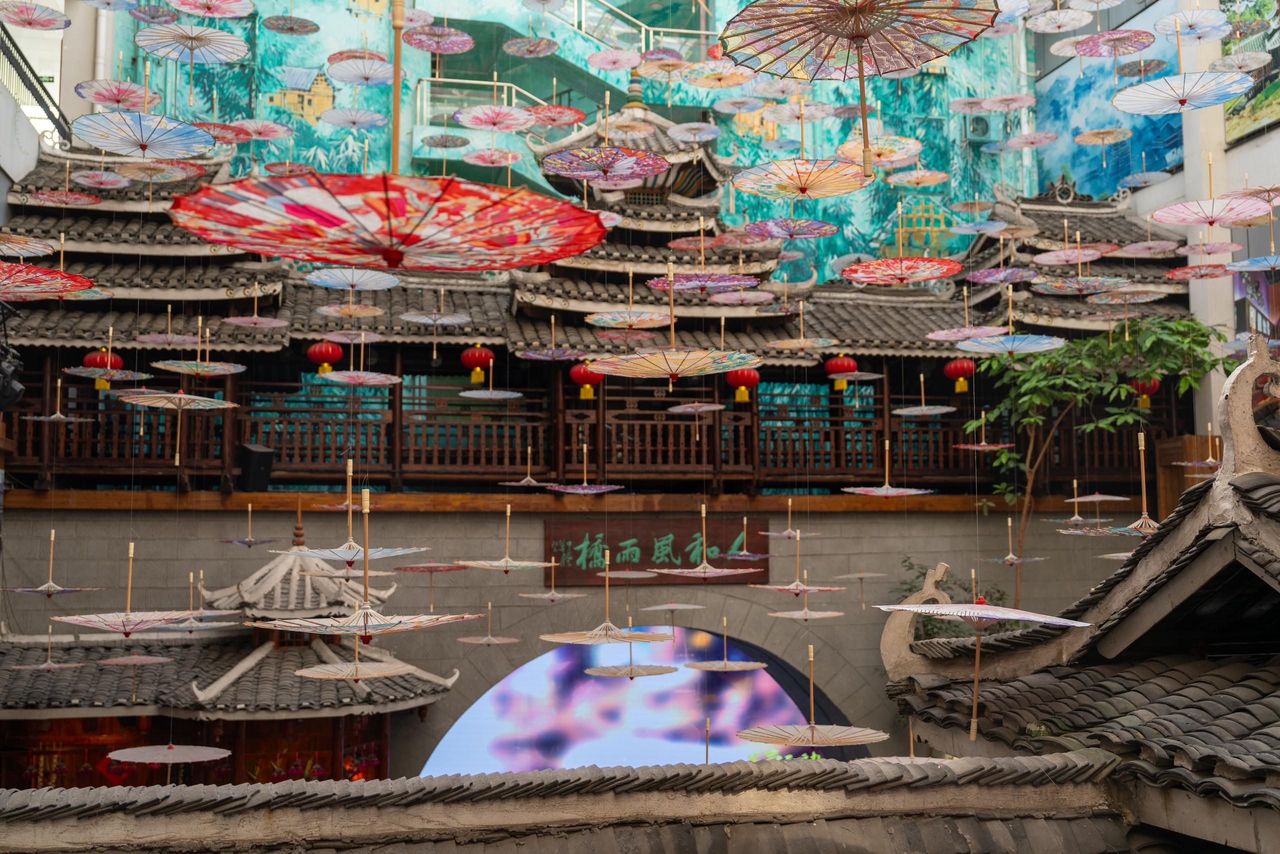 The atrium of an indoor commercial street: hundreds of painted paper umbrellas in red, pink, blue, white and other colors are hung in the space, densely covering the top of the entire frame; the background is an antique Chinese-style building, with gray tile sloping roofs, dark wooden grille doors and windows, second-story wooden railings and several red lanterns; there is a dark plaque with golden Chinese characters in the middle of the lower part of the frame, and underneath it is a circular light-emitting device.