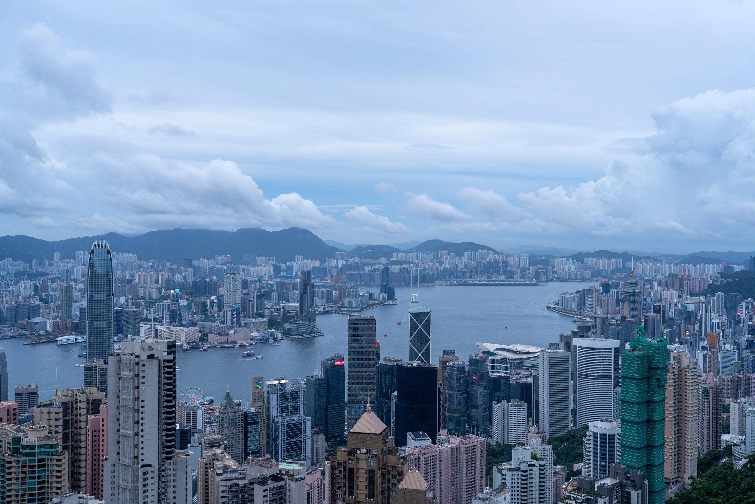A panoramic view of a port city viewed from a high place under a cloudy sky: in the middle of the frame is a wide harbor with several ships on the sea; near the shore is a large area of ​​extremely dense high-rise buildings, with a high-rise building with an obvious cross-steel frame pattern in the middle of the glass curtain wall building group; on the other side there are also high-rise buildings and neat buildings, behind it is a continuous green mountain range, and the sky is thick with clouds.