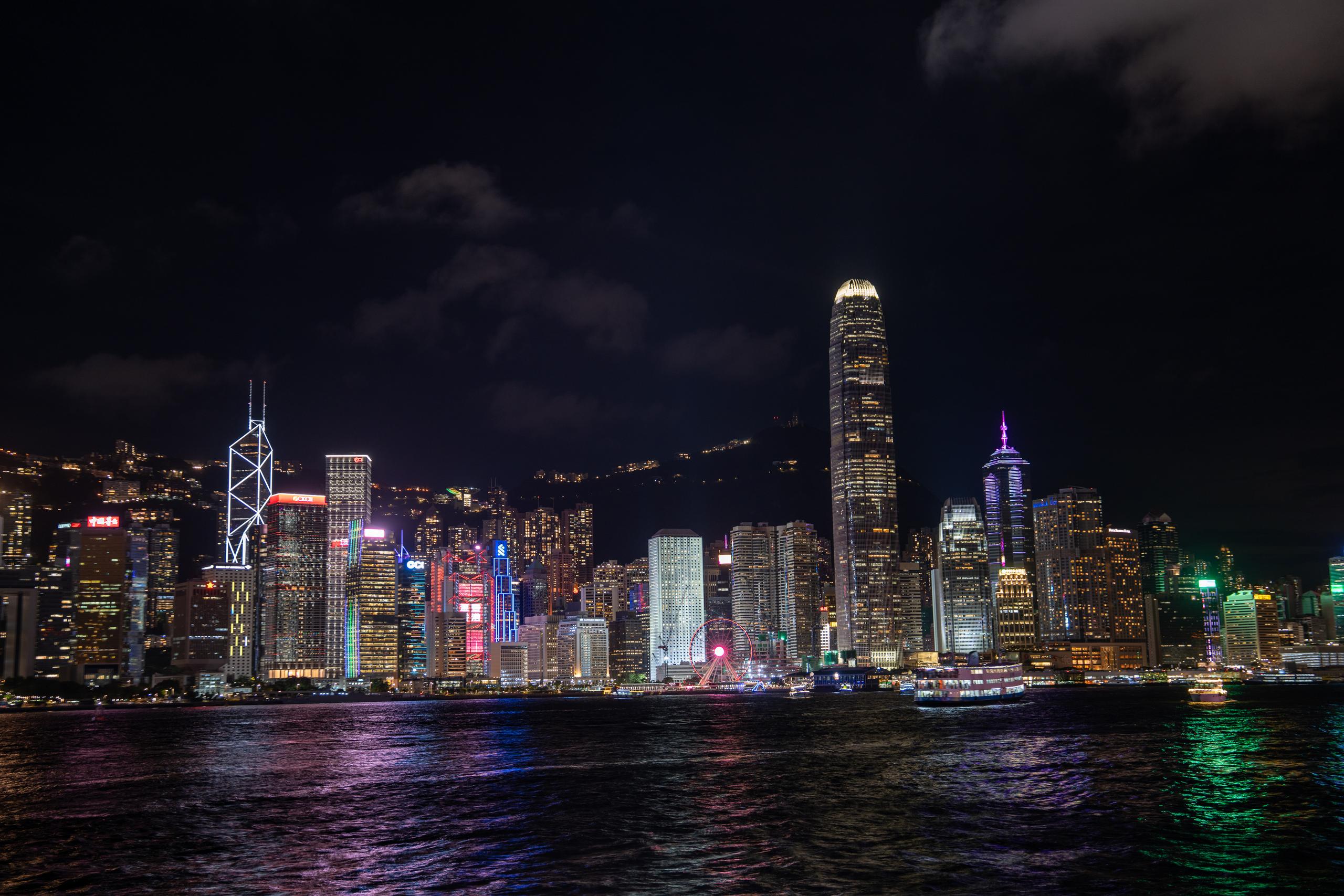 The harbor city skyline at night: dense skyscrapers are lit up in the night, mostly with white and warm yellow window lights. Several buildings have pink, blue, and green colored lights. In the middle right of the frame, there is a super high-rise cylindrical building that is illuminated as a whole; behind the buildings are dark mountain shadows, and there is a Ferris wheel with colored lights nearby; in front of the broad sea reflected the colored lights of the city.