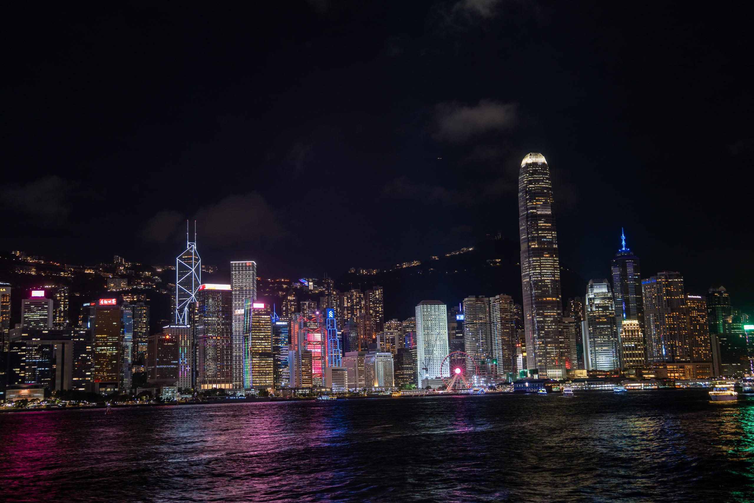 The night view of the harbor, the composition is slightly wider: the high-rise buildings on both sides of the strait are brightly lit in the night, the super-tall cylindrical building on the right is particularly eye-catching, and there is a high-rise building with an obvious cross steel frame pattern on the left; behind the buildings on the other side is the dark outline of the mountain, with a Ferris wheel with colored lights in the middle in front of the buildings; there are several cruise ships lying across the sea, and the water is full of colorful light reflections.