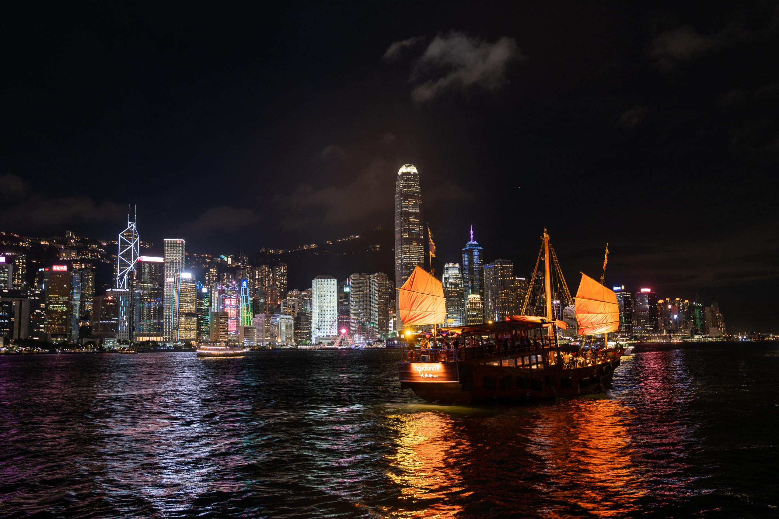 Another angle of the same harbor under the night scene: a traditional Chinese sailing ship is sailing on the water, with two orange-red sails illuminated by lights; in the background is a group of modern skyscrapers full of lights, with a tall cylindrical building to the left of the center, and a Ferris wheel with colored lights can be seen in the middle; the water in the foreground is full of ripples, reflecting orange-red and purple lights.