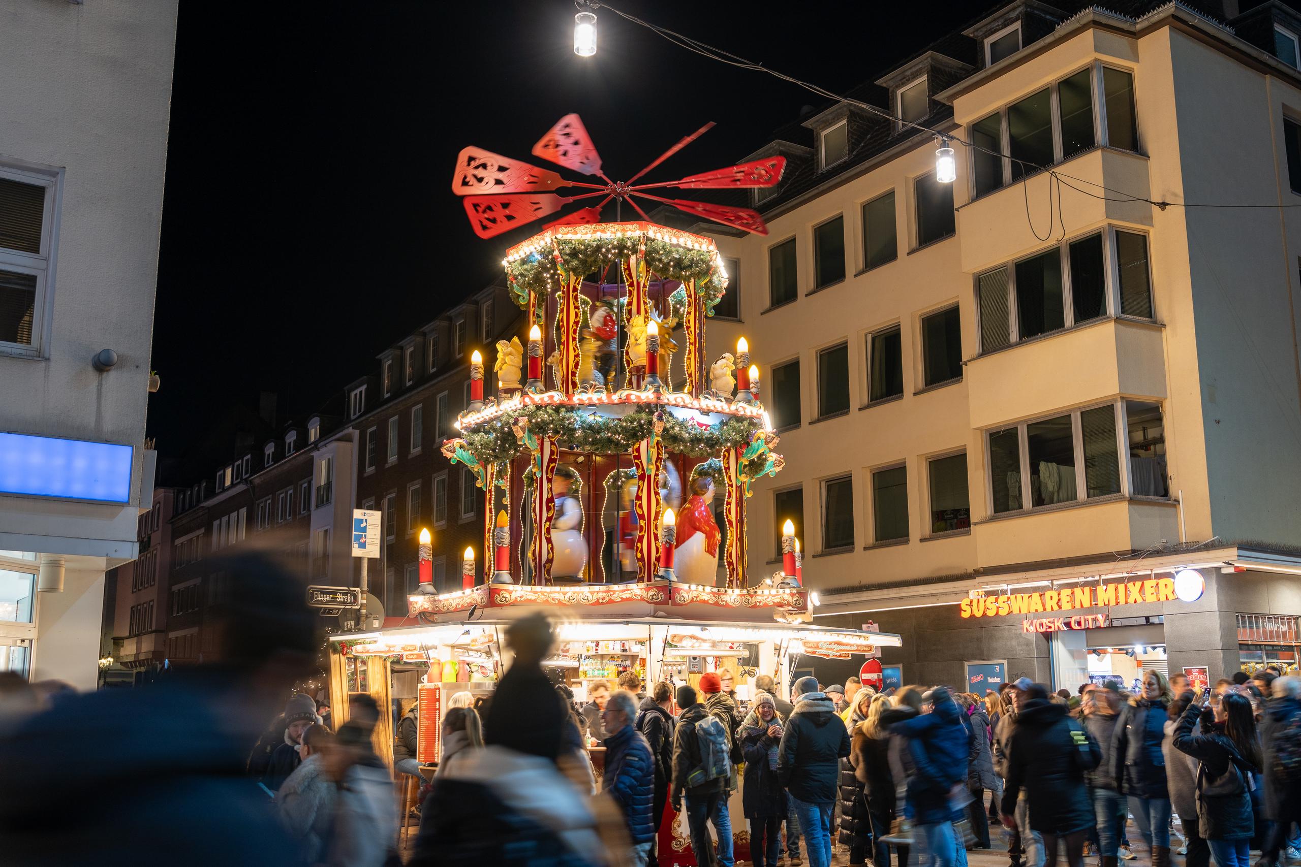 Christmas market at night: In the center of the frame is a tall carousel-style lighting installation with a red pinwheel-shaped wheel on the top. The lower part is divided into two floors. Each floor is surrounded by green garlands, gold and red lights and puppets. The installation is illuminated with warm lights. Below the installation are market stalls, crowded with tourists wearing winter clothes. The background is dark city buildings, and a shop sign on the lower right is lit with yellow light.