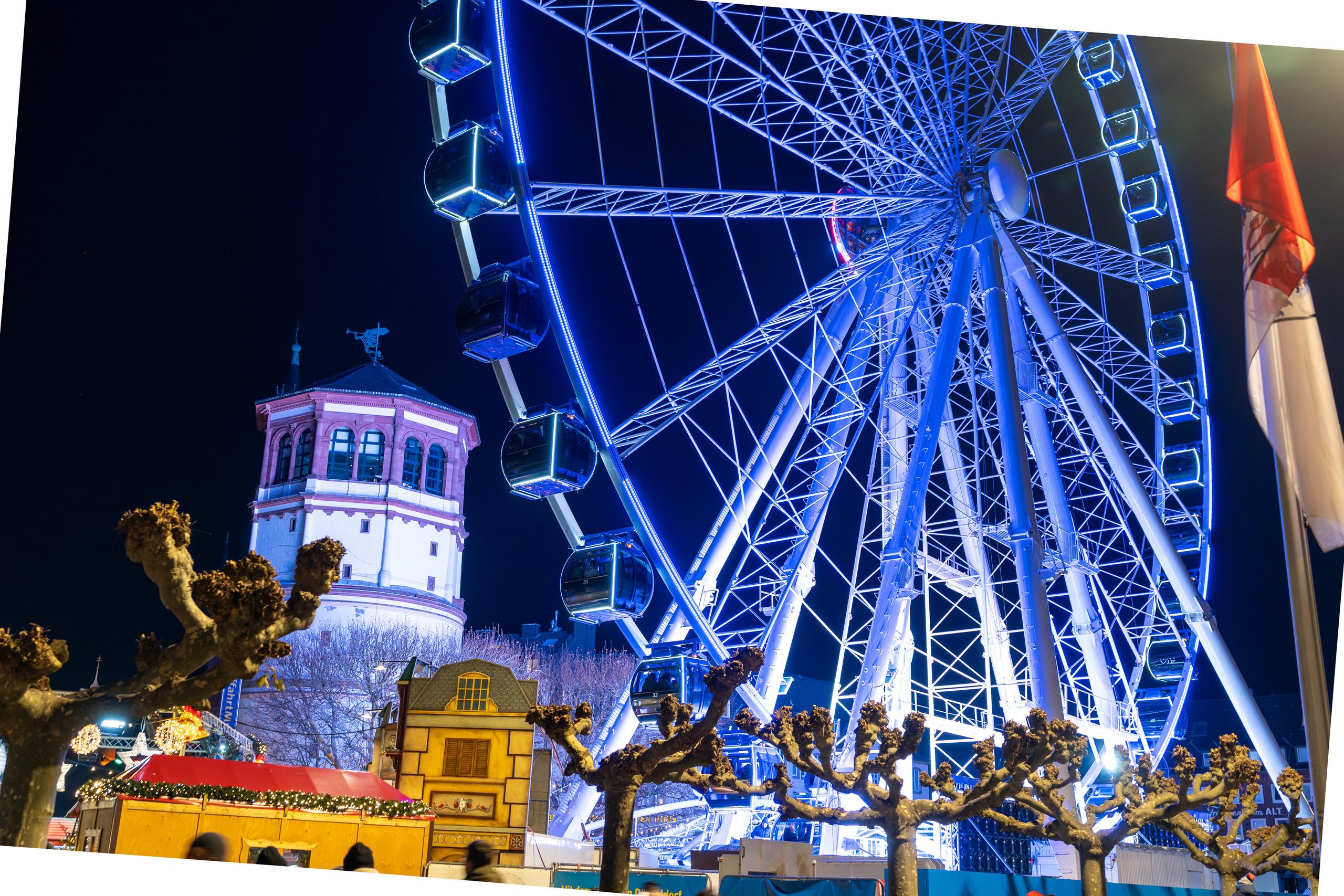 A corner of the night playground: The upper part of the frame is occupied by a huge Ferris wheel. The wheel body is composed of a white steel structure, the skeleton is illuminated by blue and white lights, and multiple square cabins are hung; on the left side of the Ferris wheel is an old-fashioned building with an octagonal tower and a red tile roof; in the foreground are several Christmas market stalls and a few bare trees trimmed into spherical branches, and the sky is dark blue.