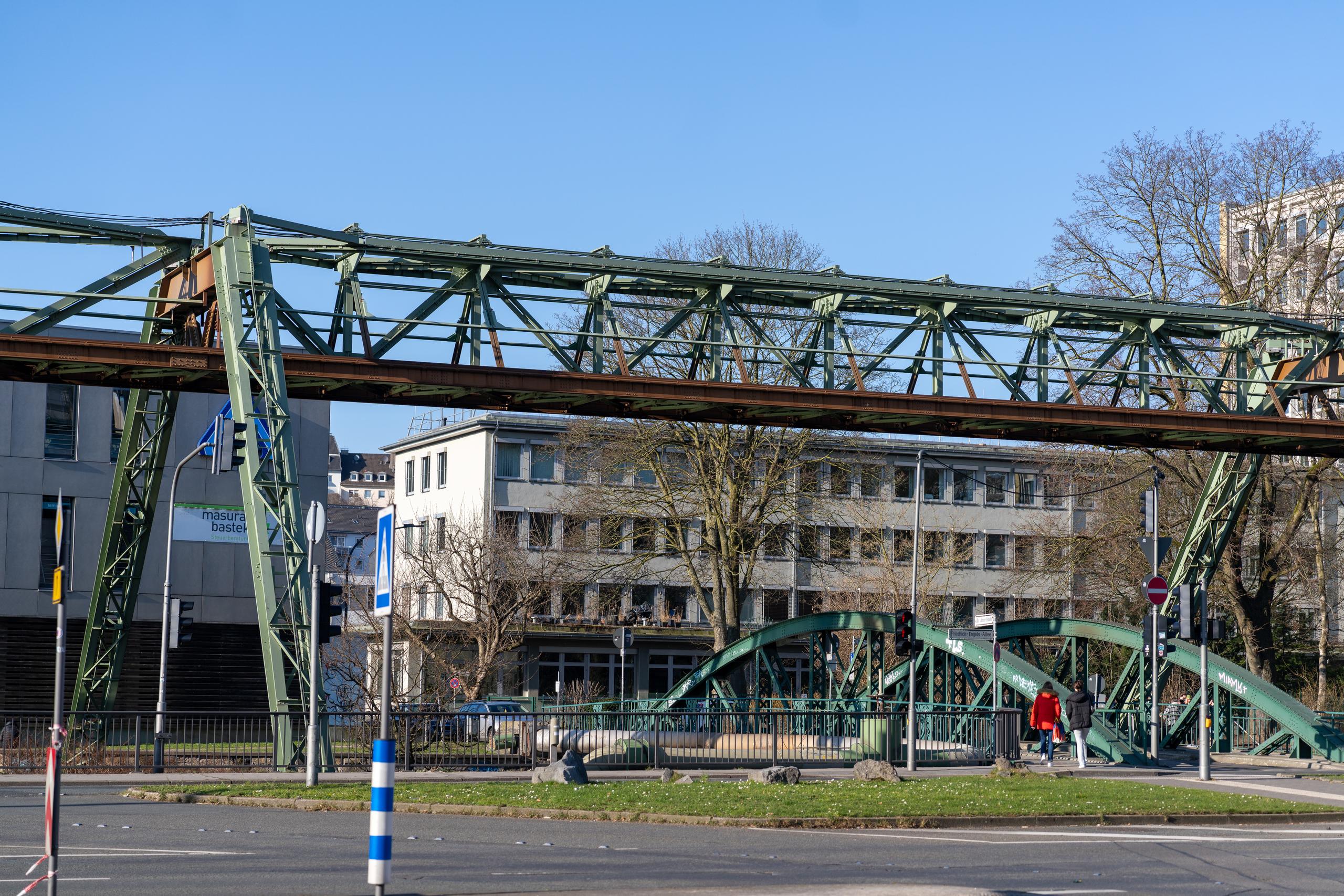 Sunny winter city street scene: A green steel structure truss bridge spans the center of the frame, with a reddish-brown bridge deck underneath; behind the bridge is a light gray multi-story building with rows of sunshades hanging under the windows; at the rear right is a green arched steel bridge and pedestrian walkway. Two pedestrians walked under the bridge. There are many traffic signs and a blue and white striped warning pole on the roadside, and the sky is blue.