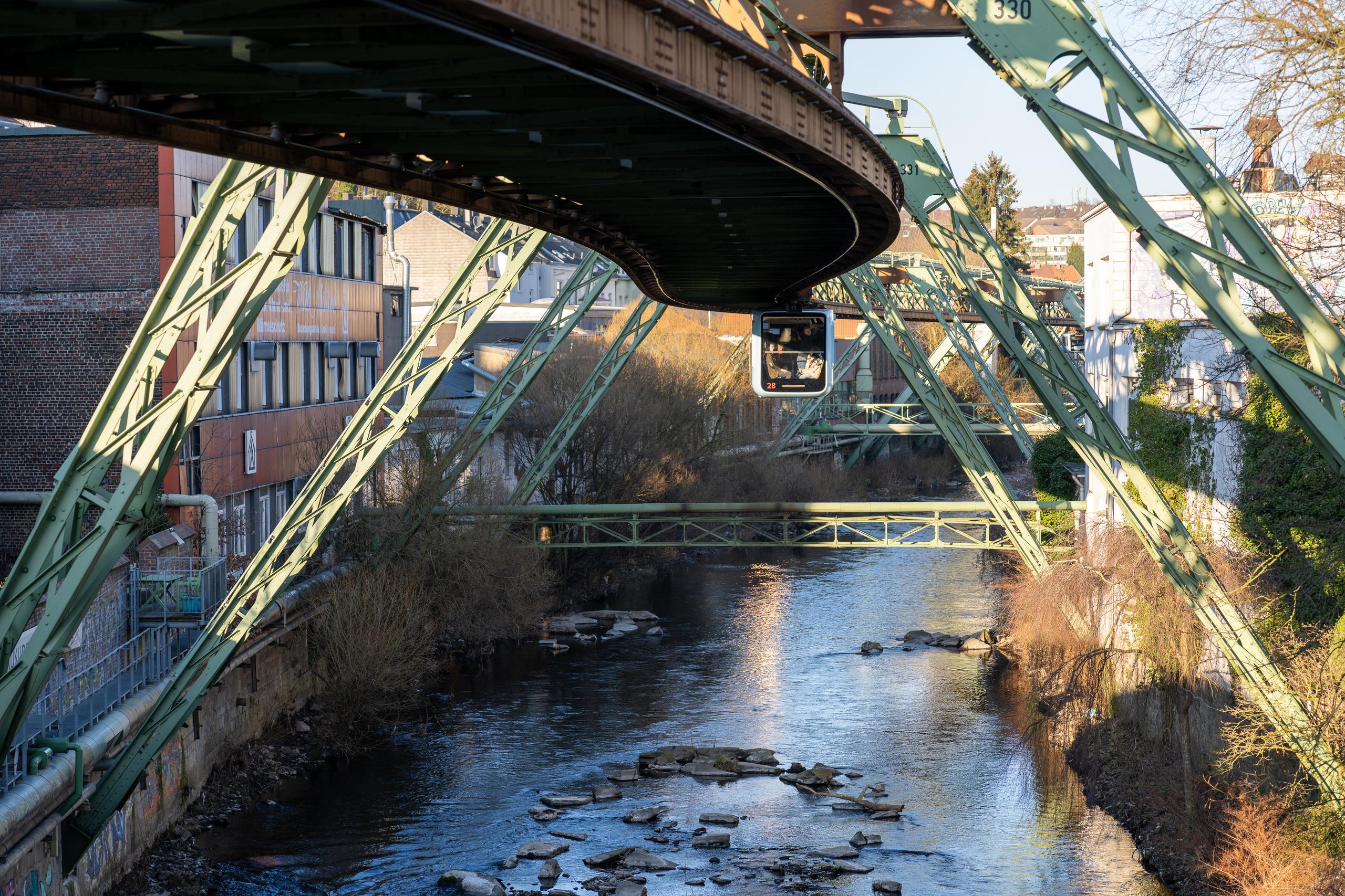 Under a green steel-framed elevated track bridge: the huge steel frame structure is gathered from the top and sides of the frame to the middle, forming a row of heavy suspended tracks, with a square rail car cabin hanging below the track; under the bridge is a shallow river, with stones and shoals scattered on the riverbed; on both sides are stone retaining walls, bare trees and red brick and white factory-style buildings, with the sun shining slantingly on the steel frame.