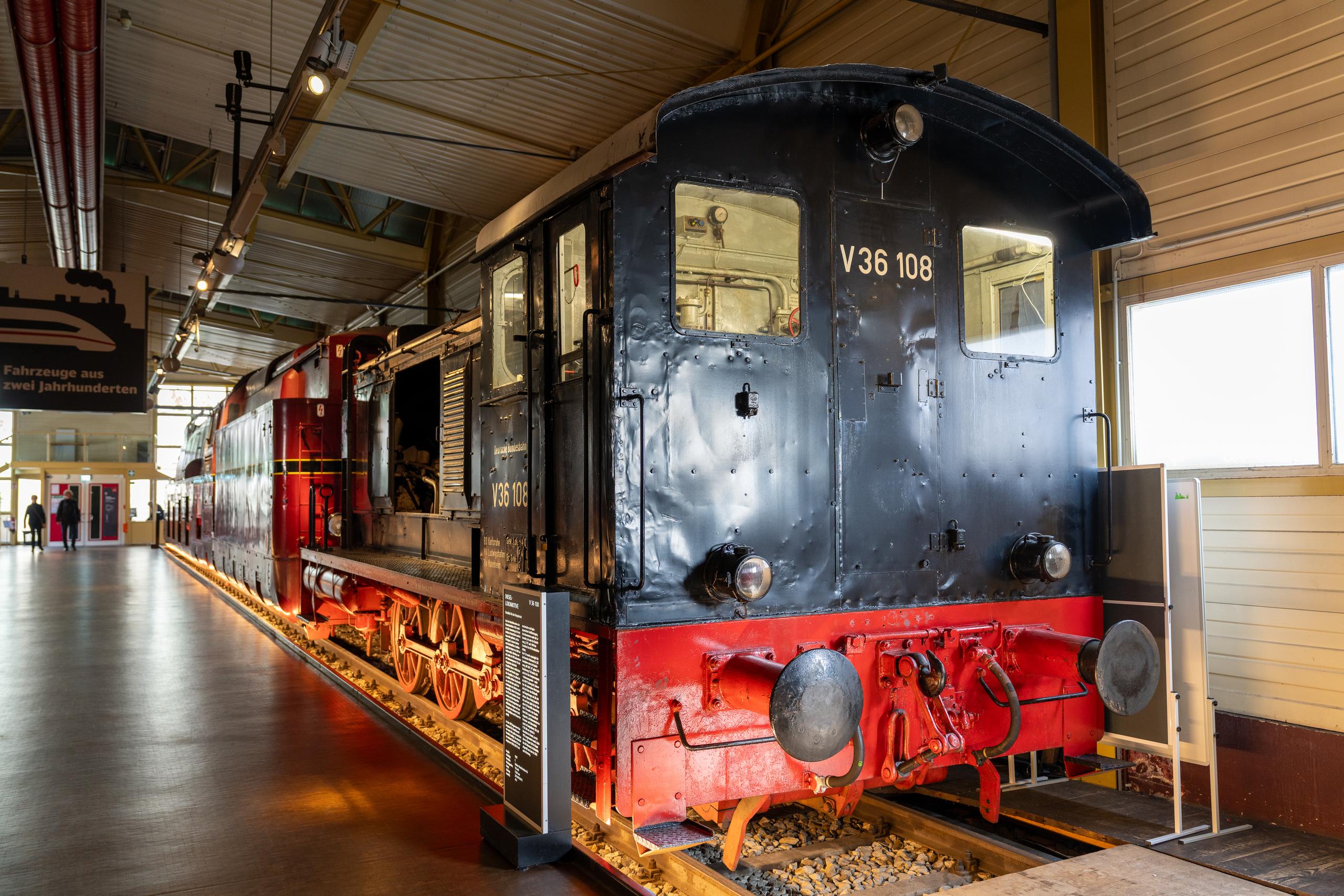 The indoor exhibition hall of the Railway Museum: The main body of the frame is an old-fashioned locomotive. The front of the locomotive is black and covered with rivets. The lower skirt is bright red. There are two circular buffers and lights in front of the locomotive. Red wheels and connecting rod structures can be seen on the side of the locomotive. A black information board stands next to the locomotive. On the left is a long exhibition hall passage, with the backs of two visitors in the distance.