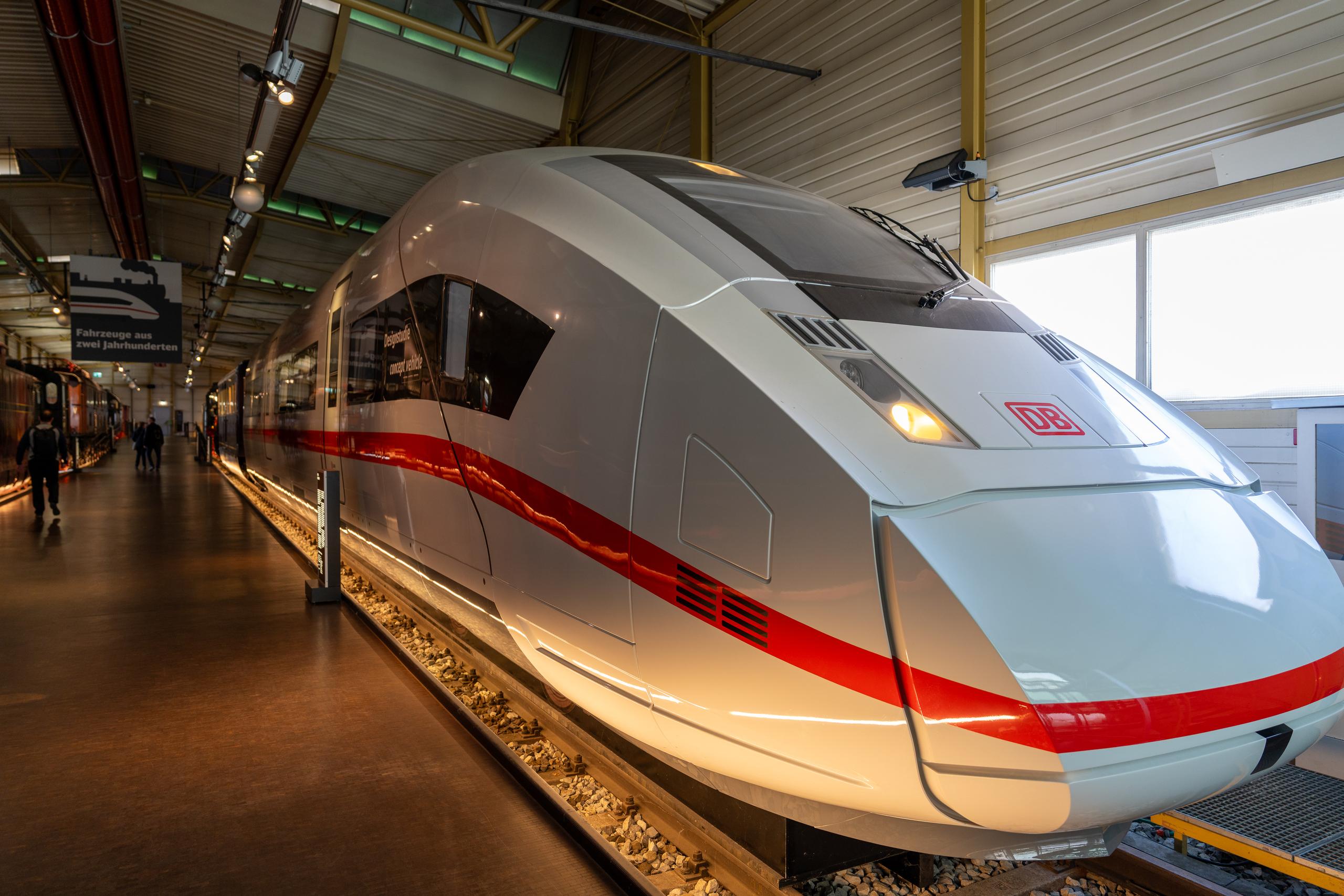 A train in the exhibition hall of the Railway Museum: the streamlined front of a modern high-speed train, with a white body and a bright red horizontal strip. The cab has a dark windshield and glass side windows. There is a red sign and two bright lights on the front. The train is parked on a gravel-paved display track. On the left is a long dark reflective ground passage. Other carriages on display can be seen in the distance.
