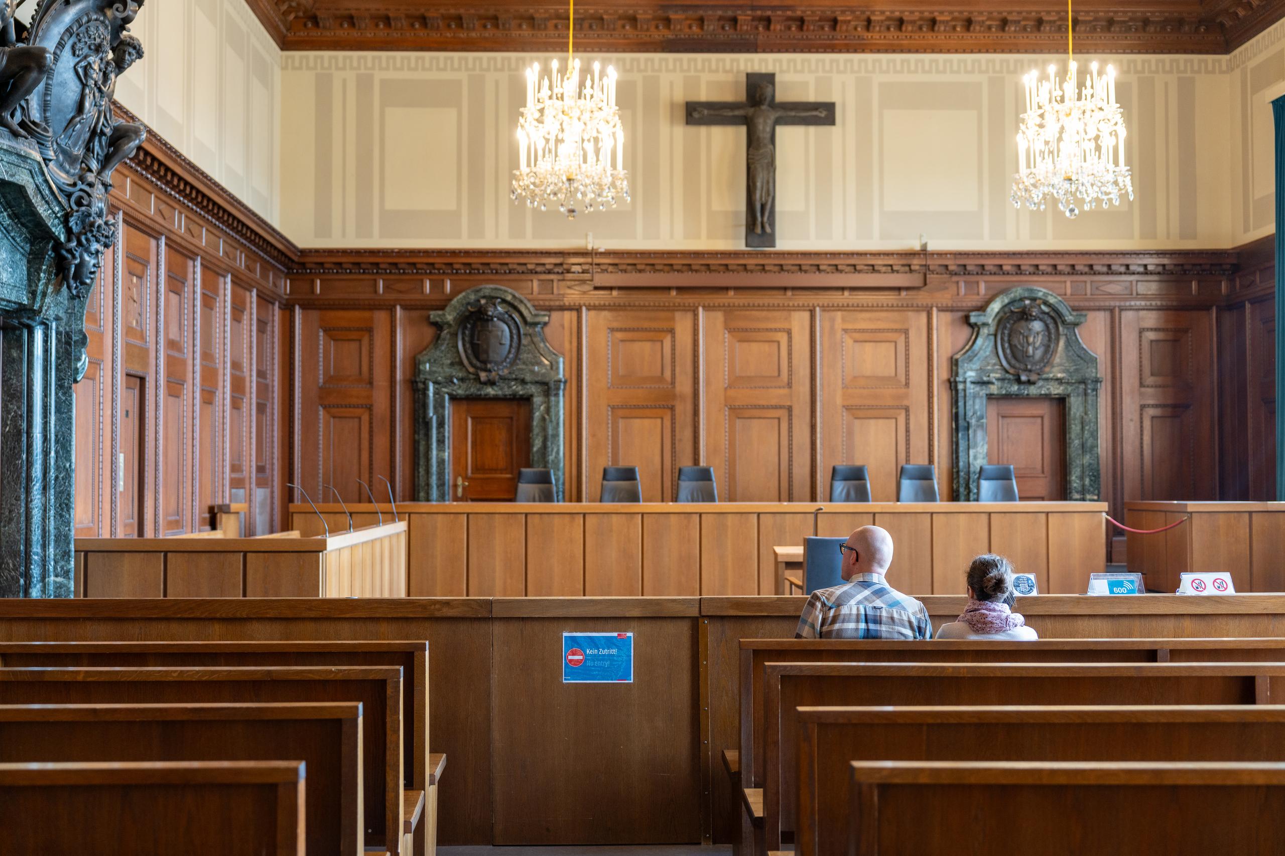 A historical court room: in the front is a dark brown wooden wainscot, with a dark wooden cross hanging in the center above the wall; two crystal chandeliers hang from the ceiling; in front of the wall is a row of high-backed dark seats and a long judge's desk; in the foreground are rows of wooden benches, with two people sitting in the second row facing away from the camera.
