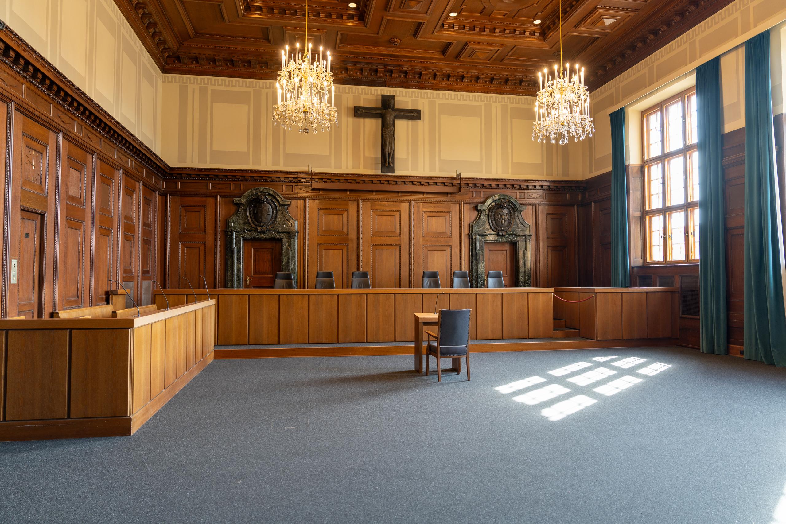 A historical court or meeting hall: on the left and directly in front is a dark brown wooden wainscot; a dark wooden cross hangs in the center of the high wall, and two crystal chandeliers hang from the ceiling; in front of the wall is a row of dark seats and a long wooden railing; on the right, a large window with a square window is hung with long dark green curtains, and the sunlight casts square spots of light on the ground.