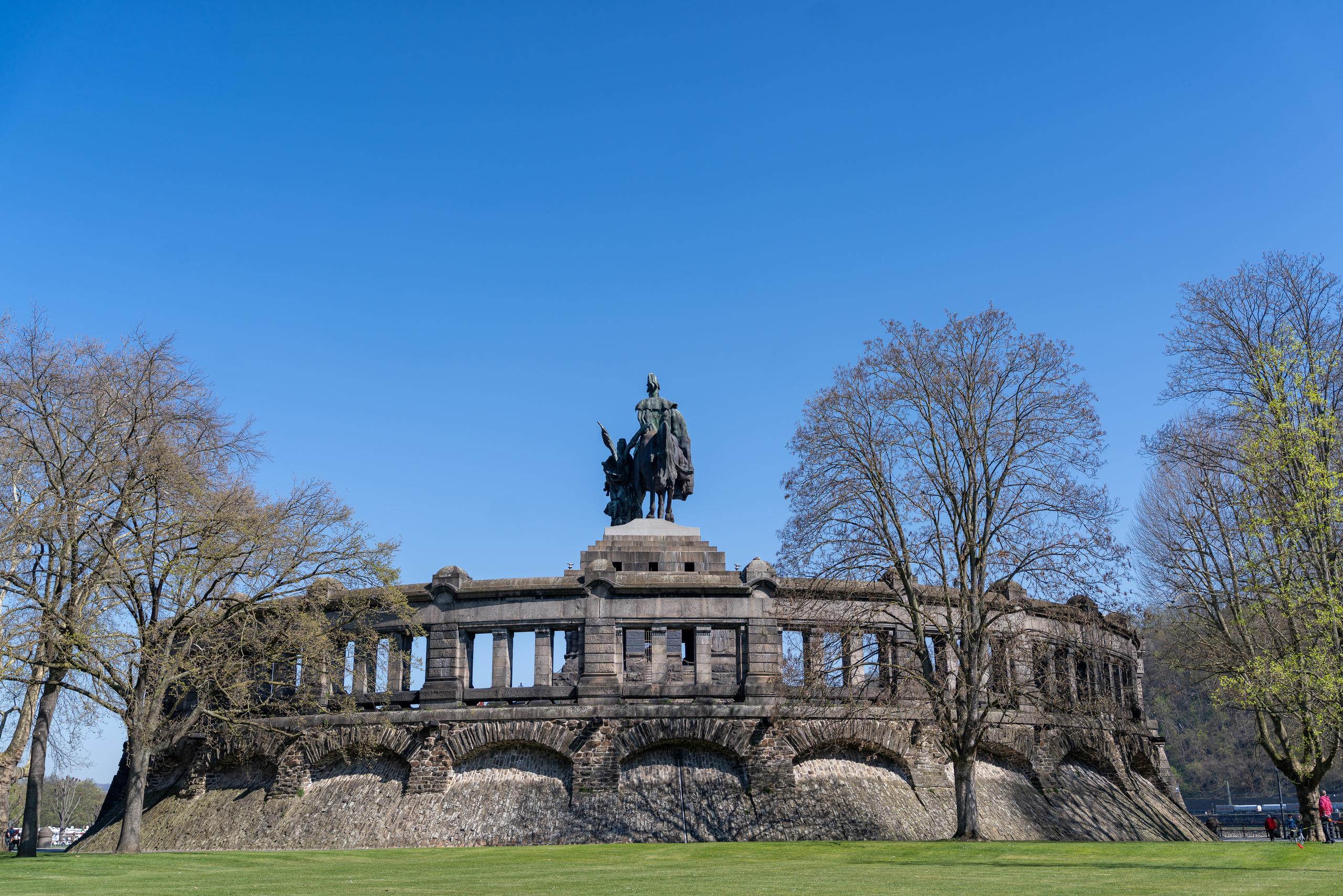 A magnificent stone monument under the clear blue sky: the monument is a semicircular stone building, with a heavy base with arched niches on the lower floor, and a column structure with square openings on the upper floor; a statue of an equestrian figure stands on the top of the monument; there is a large tree with almost bare branches on each side of the monument, and a new green grassland is on the lower left, and the sky is pure dark blue.