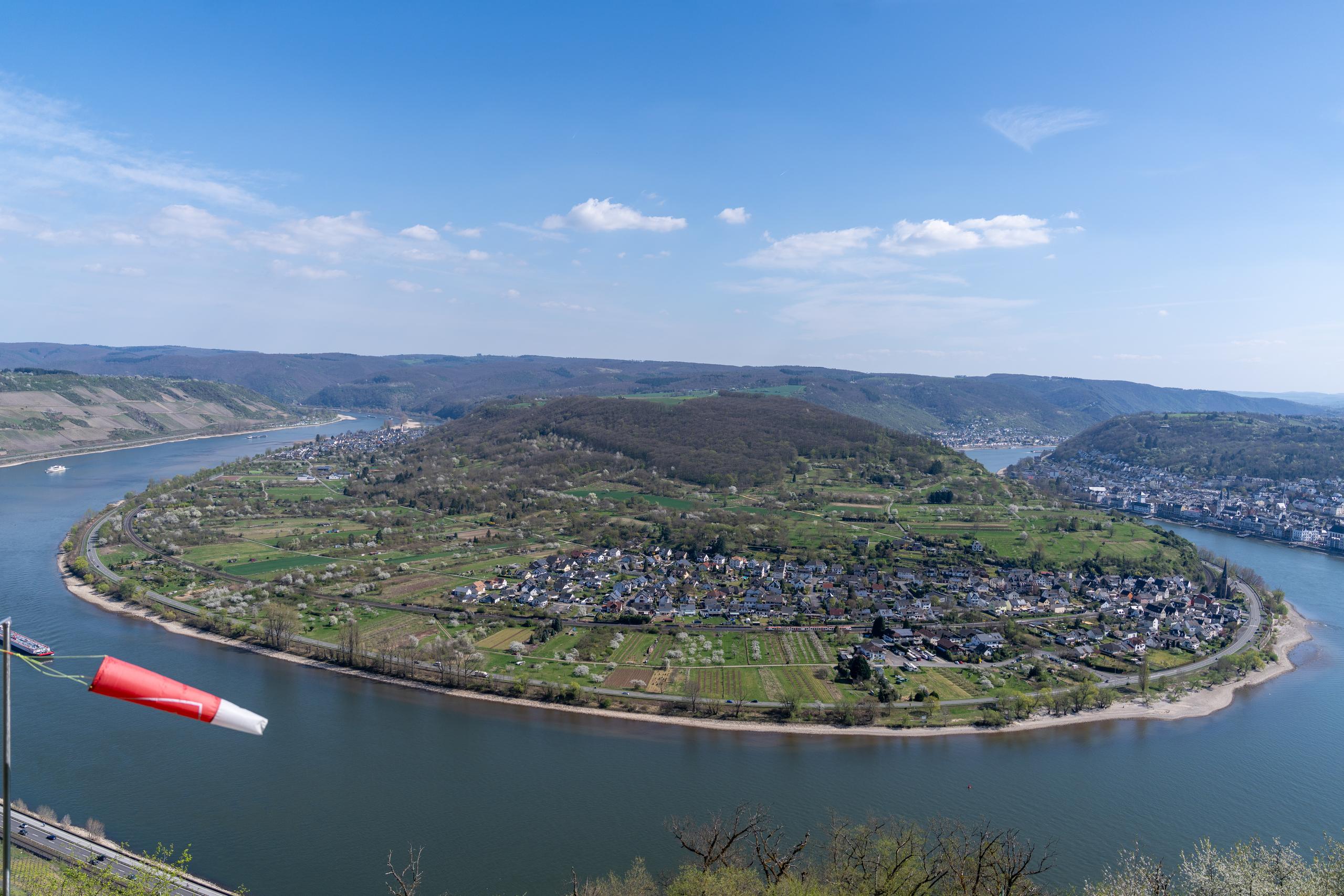 A panoramic view of the valley peninsula seen from a height: a wide river bends into a huge horseshoe shape in the frame, with a flat peninsula surrounded by the river in the center; on the peninsula are large ridges, vegetable fields, and groves of woods, with a village clustered in the middle, and a church among the densely packed houses with dark pitched roofs; on the other side of the river and in the distance are rolling low hills, and a barge is sailing on the river.